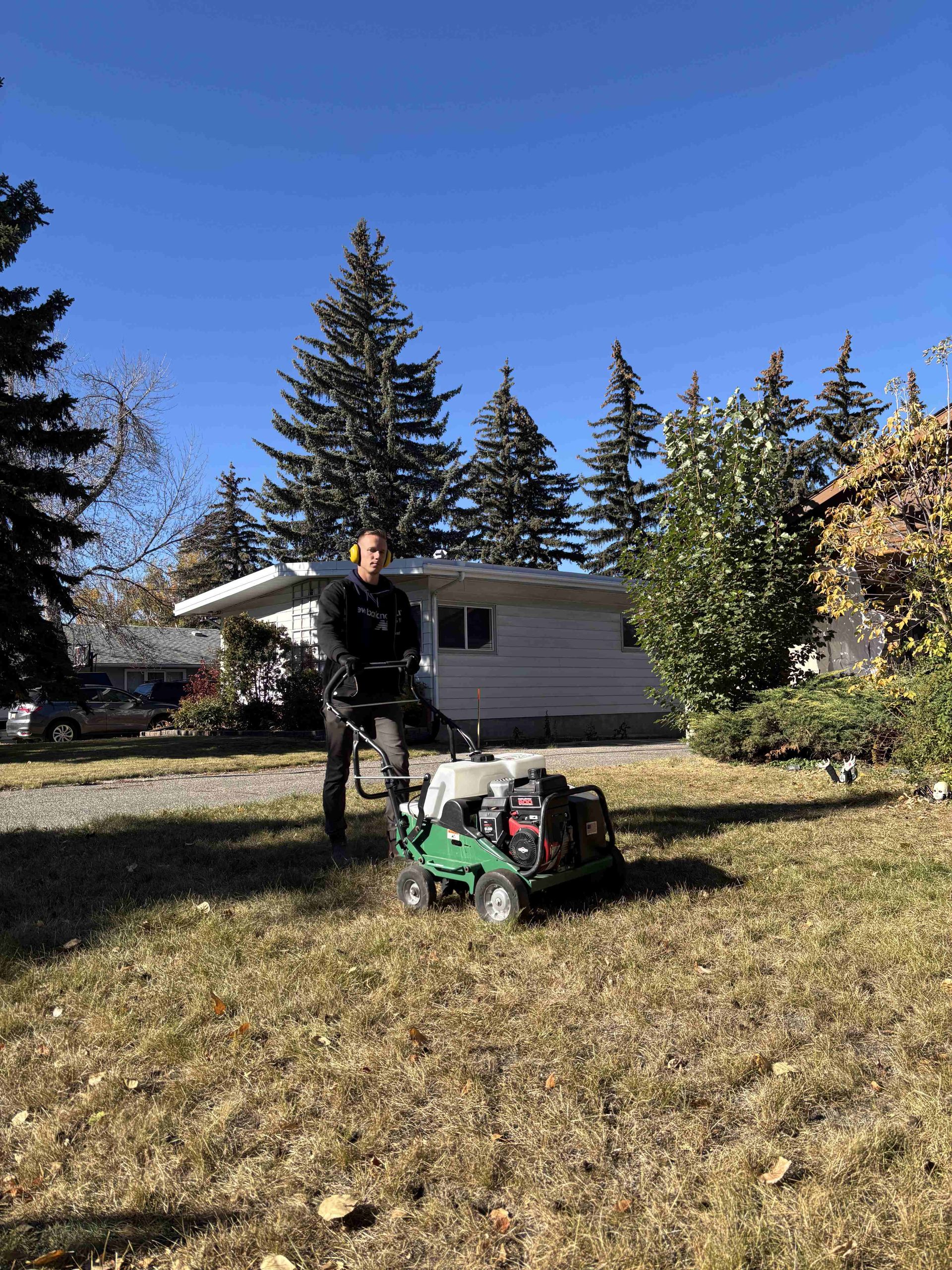 Person standing beside a lawn mower in a sunny yard with a small house and pine trees behind them