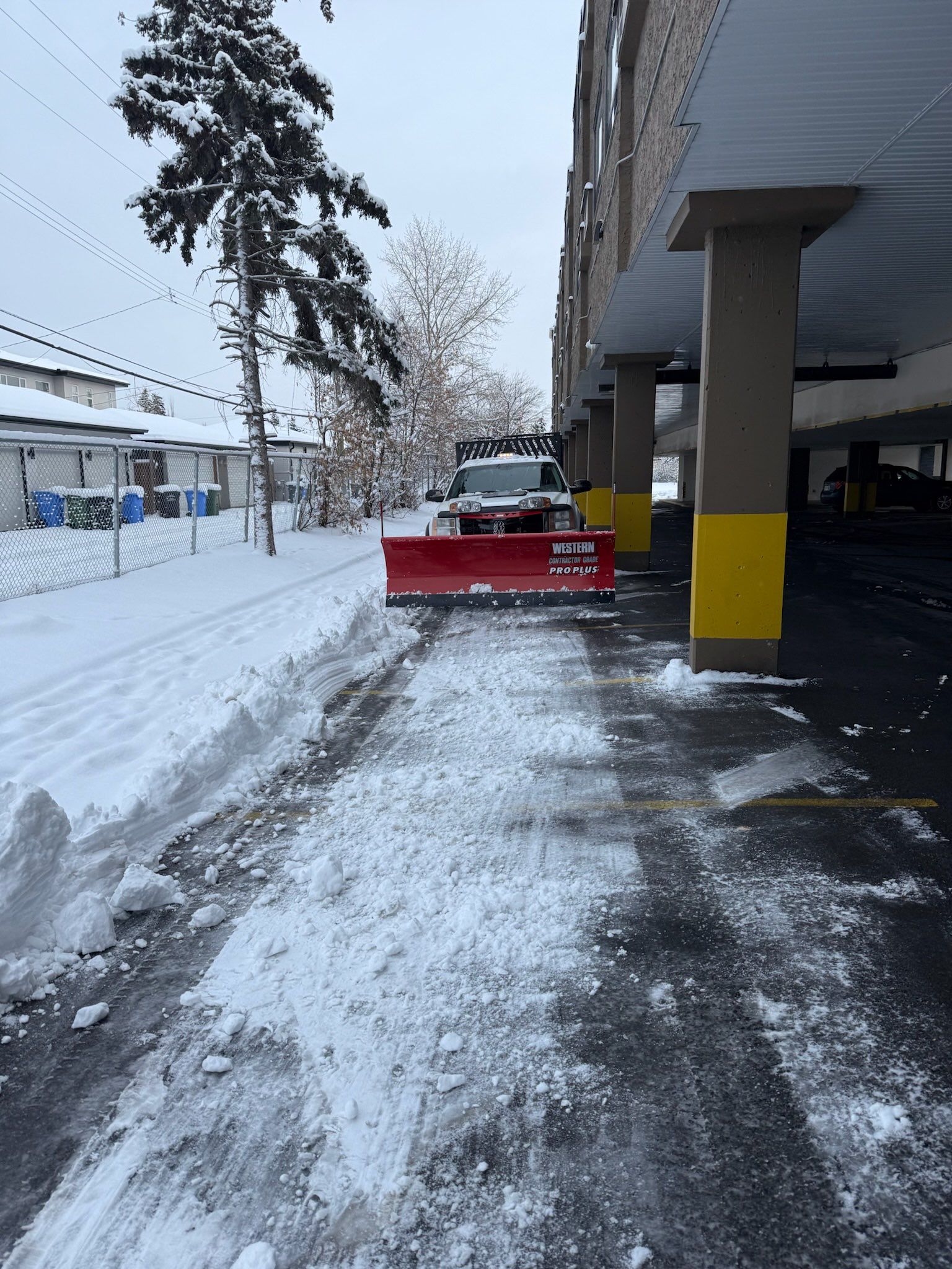 Snowplow clearing snow from a parking area next to a building. Gray sky, snow on the ground, red blade.