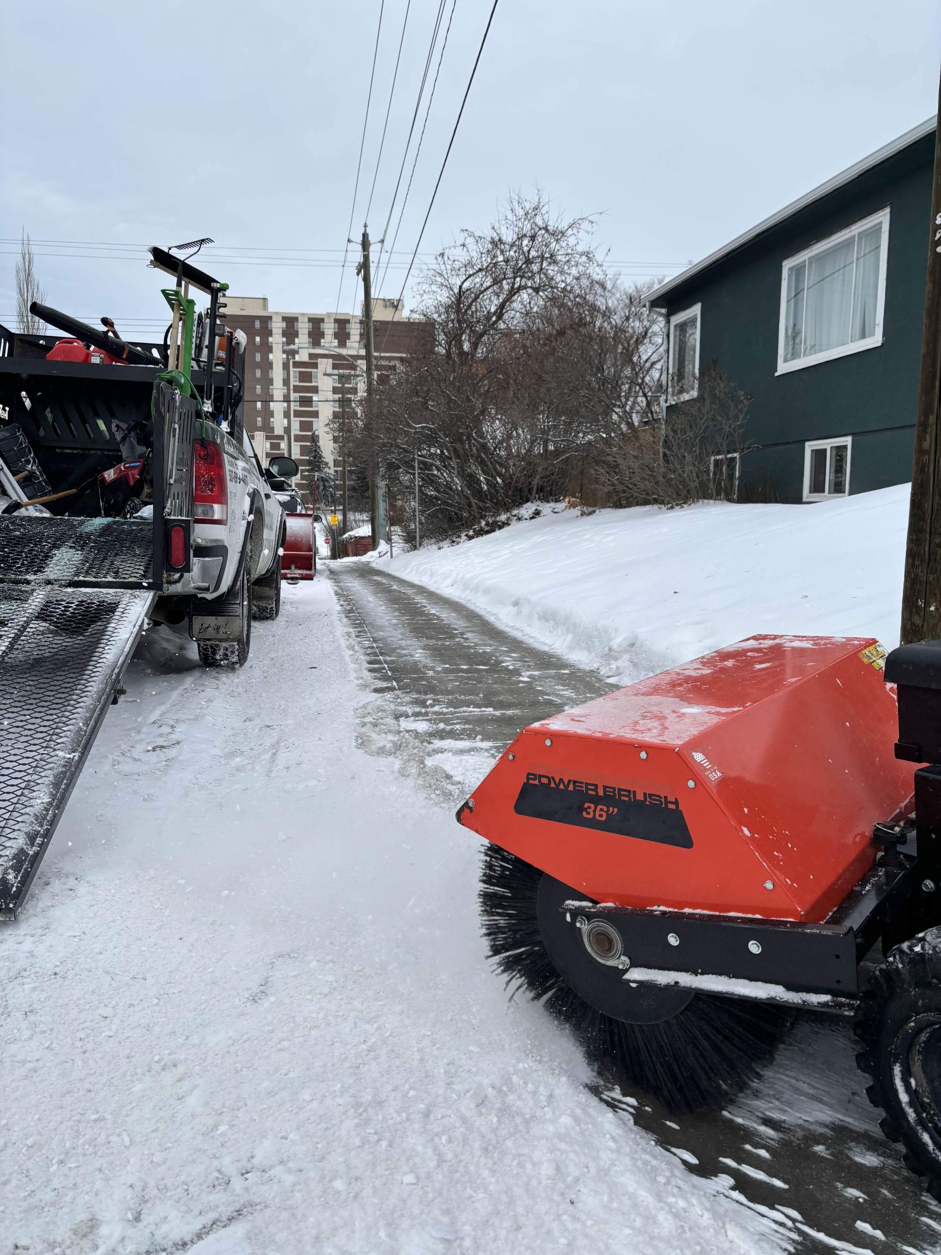 Snow sweeper clearing a snow-covered street. Truck parked alongside. Buildings and trees in background.