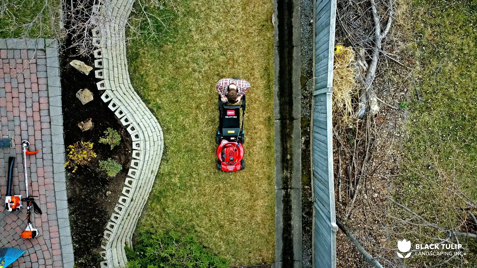 Overhead view of a person mowing a narrow lawn with a red mower, flanked by a flower bed, brick patio, and fence.