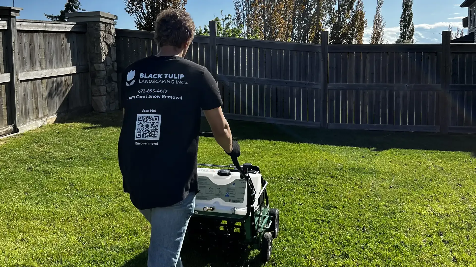 Person in black t-shirt and jeans mowing a green lawn with a white and green machine in a backyard with a wooden fence.