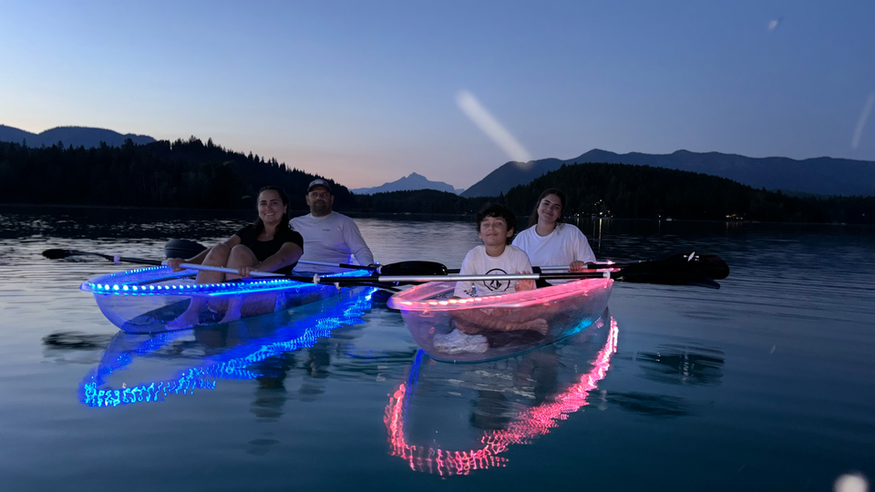 A family paddles blue and pink LED clear kayaks on calm Montana water at dusk.