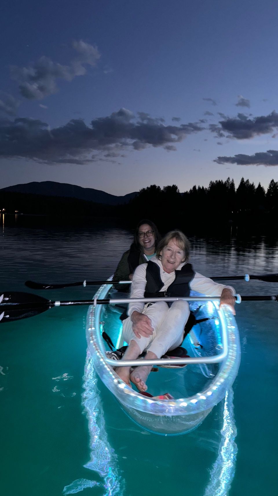 Two smiling guests sit in a clear kayak with blue LED lights at dusk.