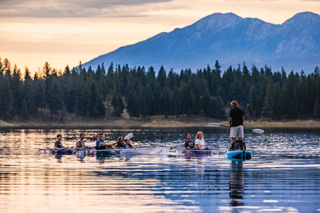 Clear kayak group on a forest-lined lake beneath Montana mountains at sunset