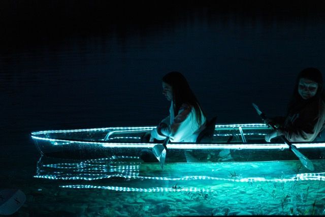 Guests in a transparent kayak illuminated turquoise on the water after dark.