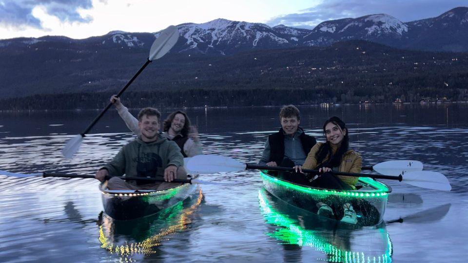 Guests paddle two glowing clear kayaks beneath mountain scenery at dusk in Whitefish.
