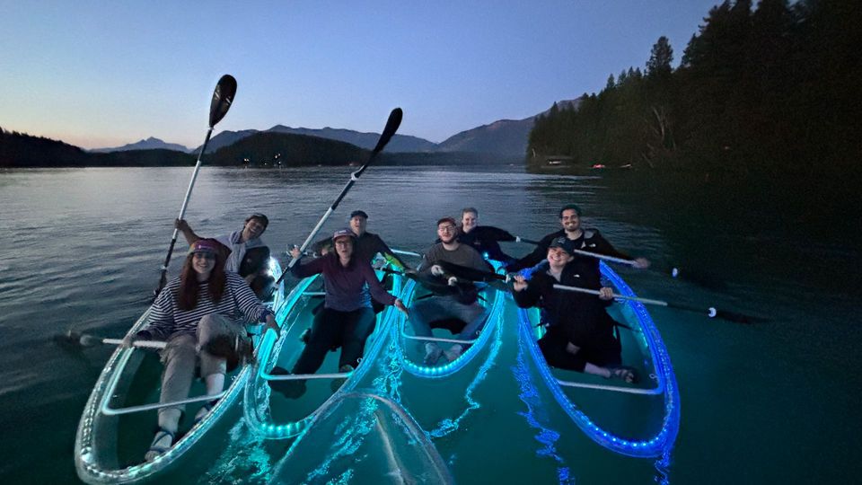 Friends gather in blue and turquoise LED clear kayaks during a Montana evening paddle.