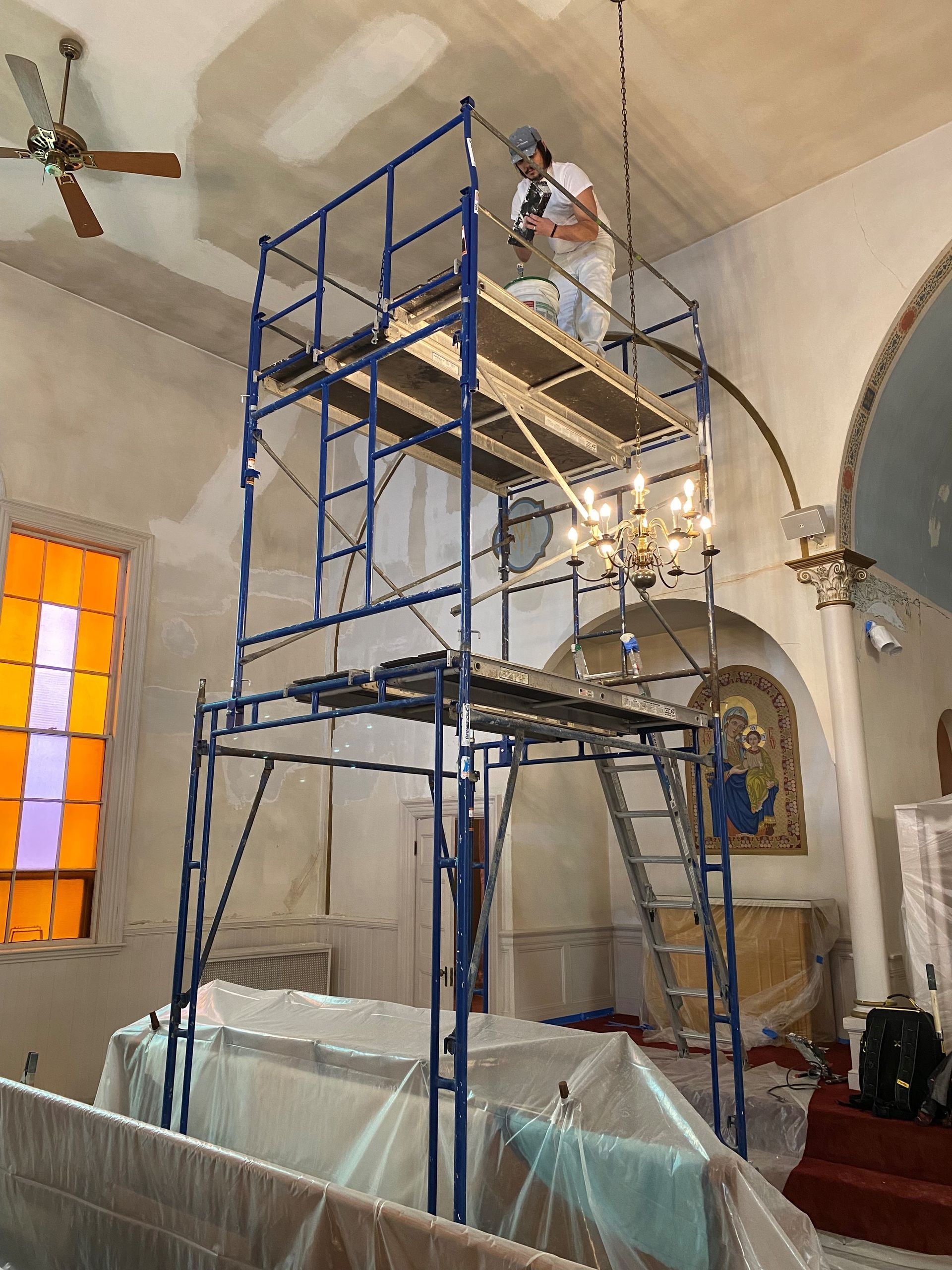 Worker on blue scaffold in church, near chandelier and stained-glass window.