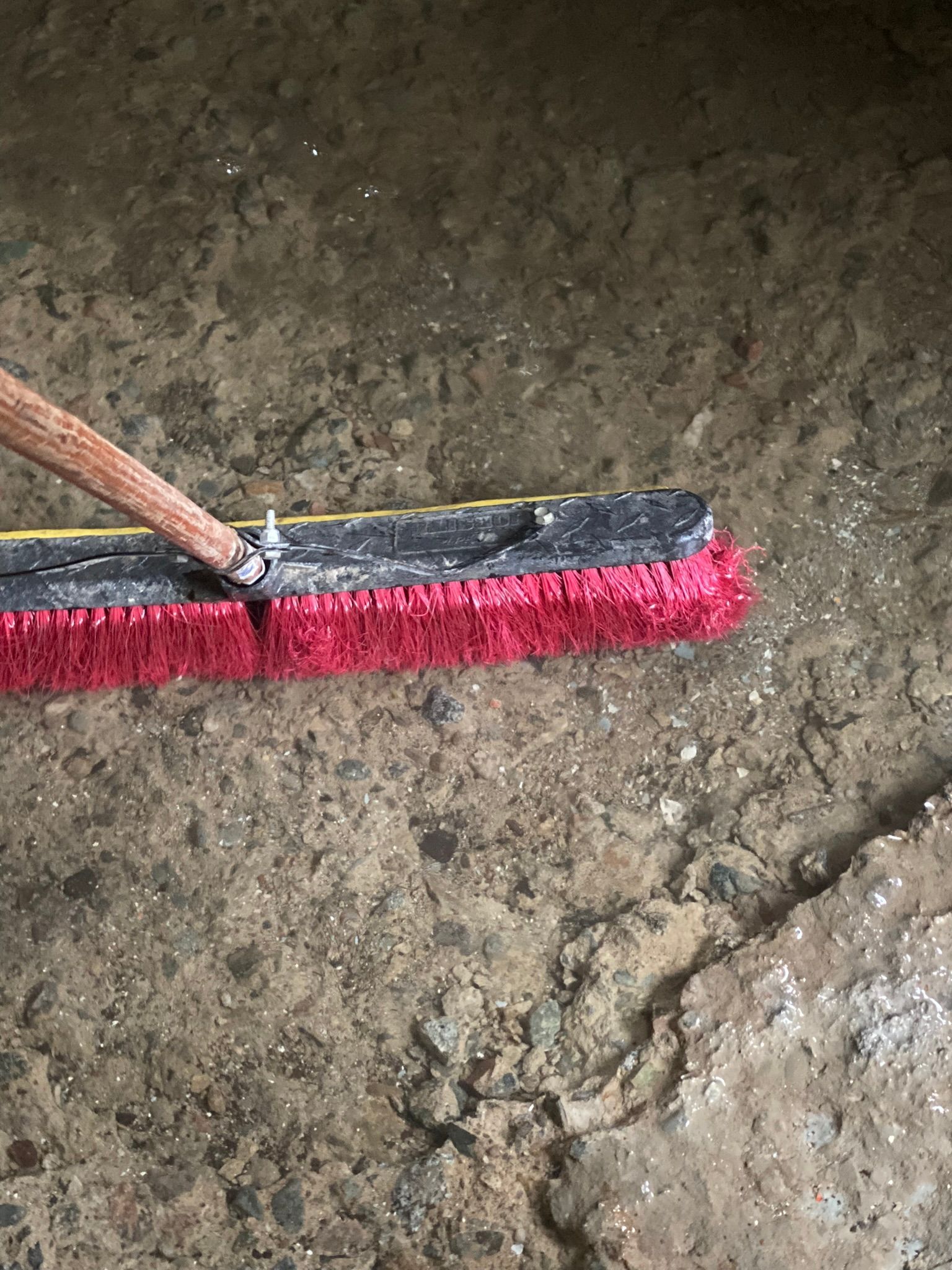 Broom cleaning dirt on a rough concrete floor, with red bristles and a wooden handle.