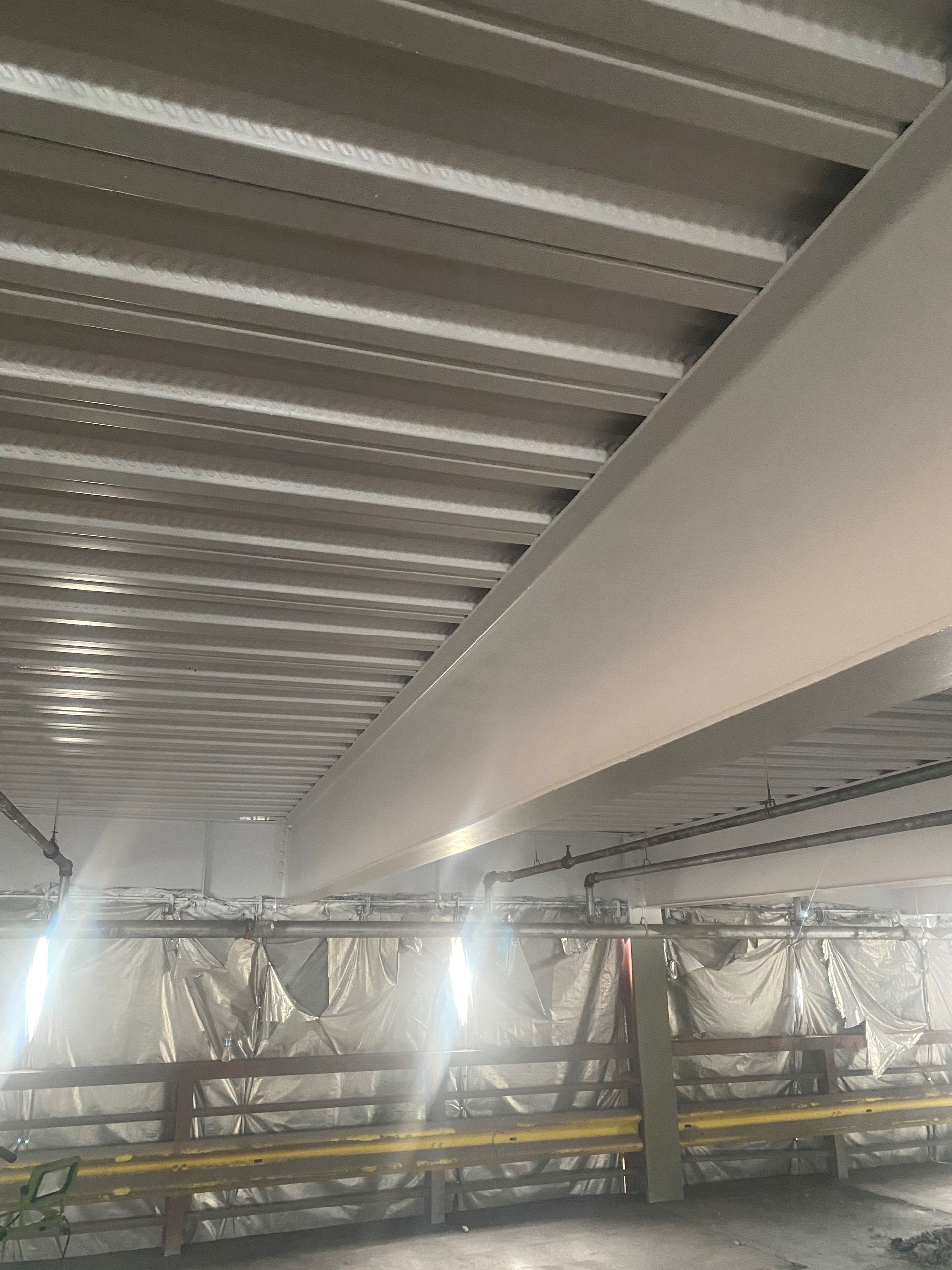 Industrial interior with metal ceiling beams, bright lights, and wrapped equipment below.