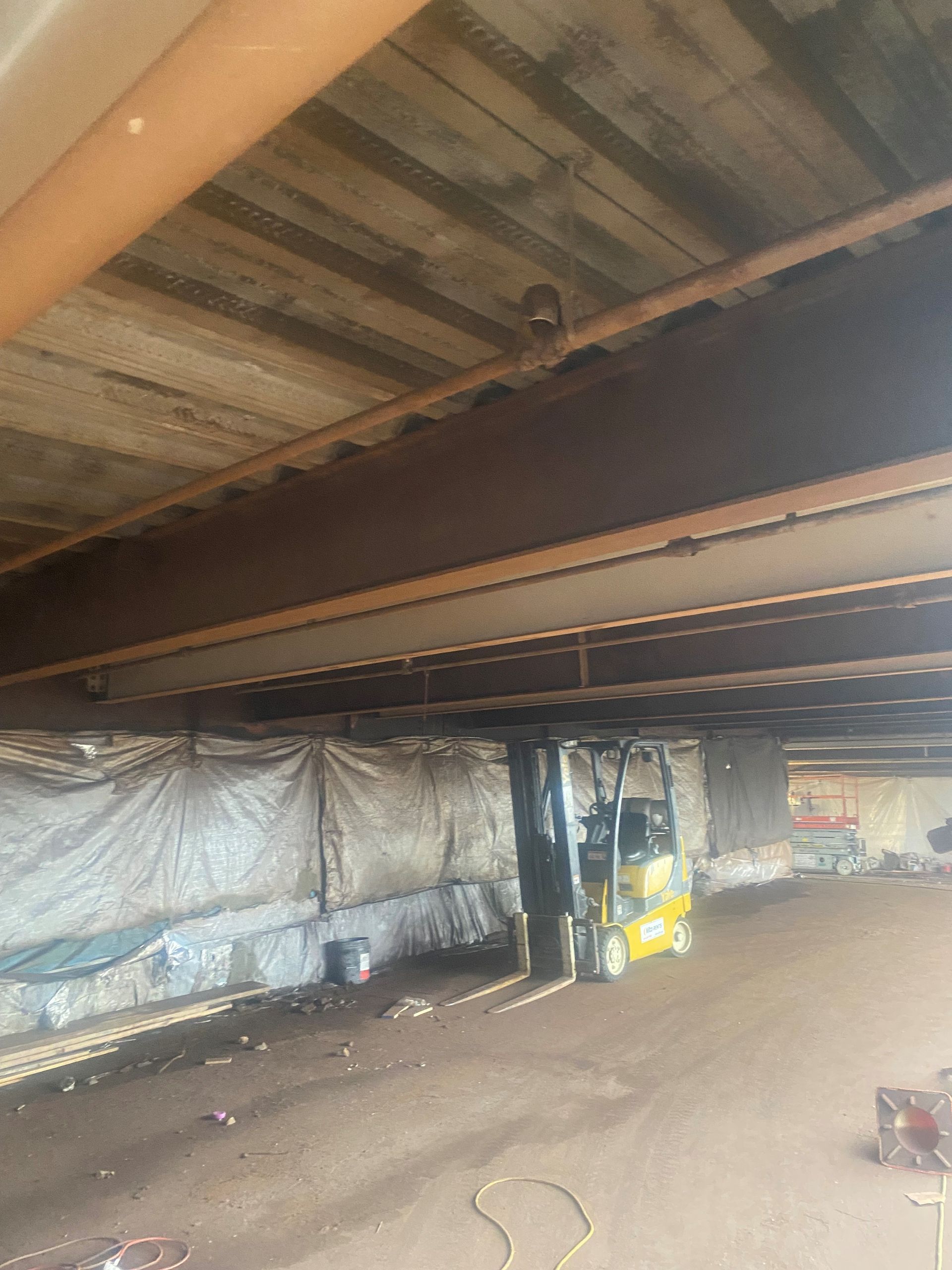 Under a metal roof, a partially unfinished interior with exposed beams, tarps, and a forklift-like machine.