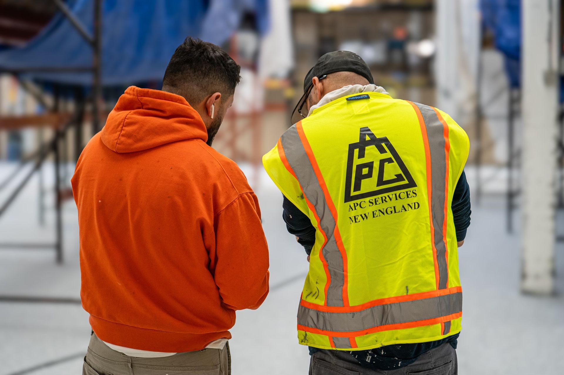 Two workers in an industrial warehouse, one in an orange hoodie and one in a reflective safety vest.