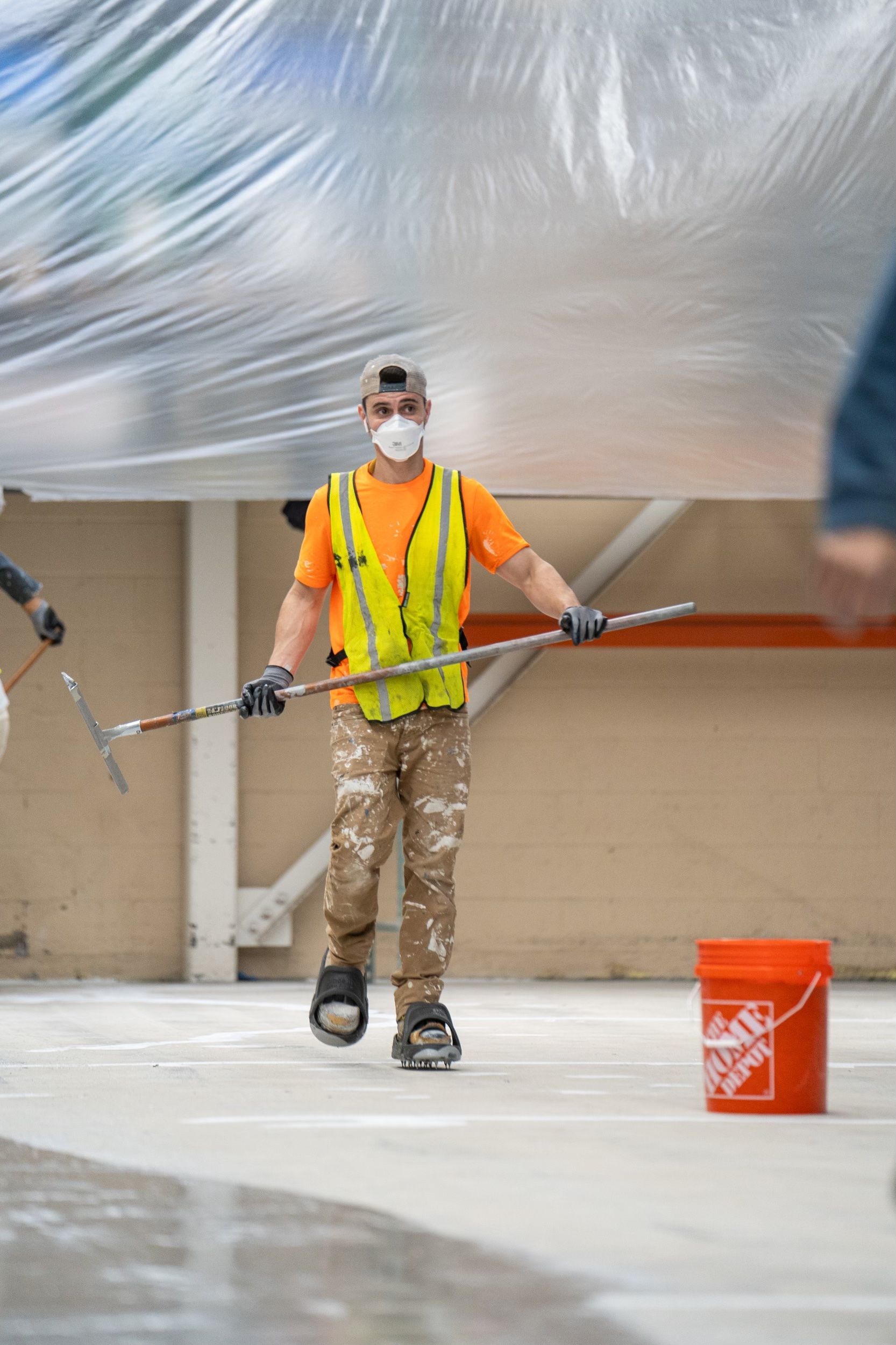 Worker in safety vest and mask painting a floor with a long-handled roller indoors