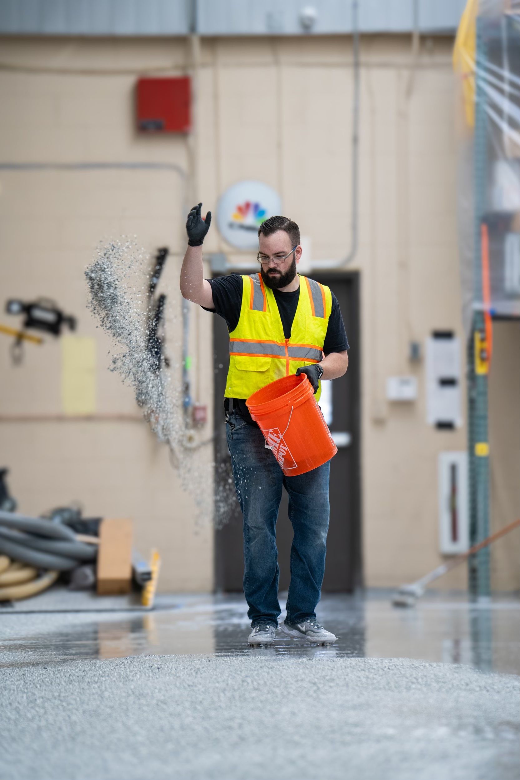 Worker in a yellow safety vest carrying an orange bucket as water splashes in an industrial workshop.