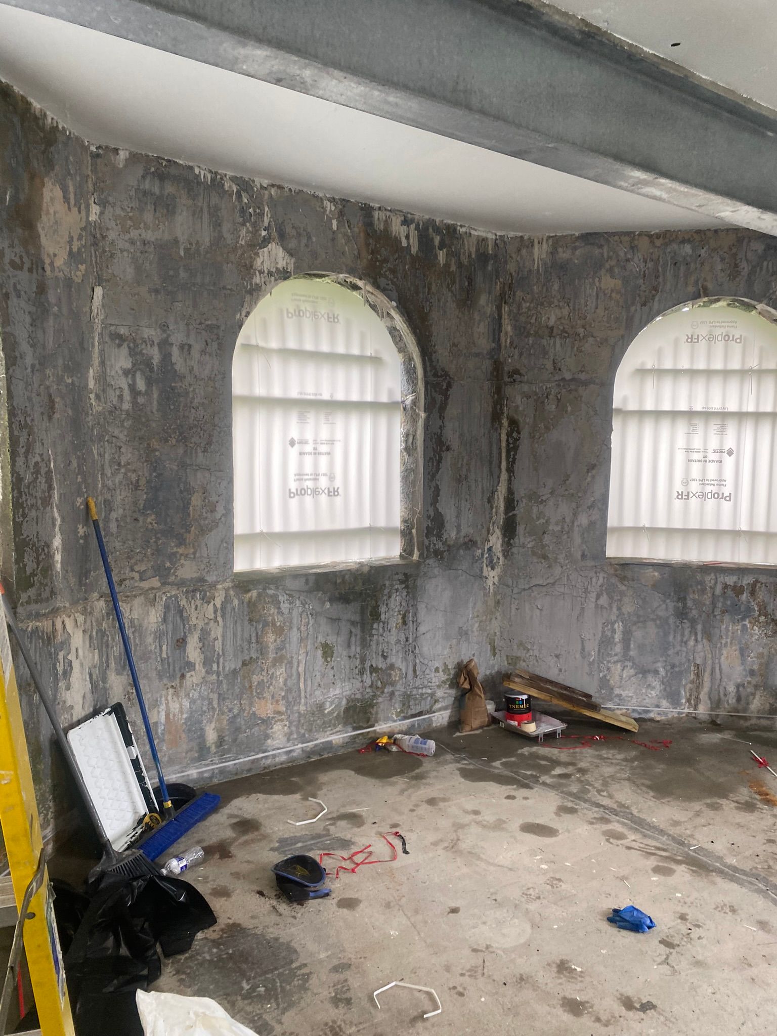 Damaged room with peeling gray walls, two arched windows, and debris scattered on the floor.