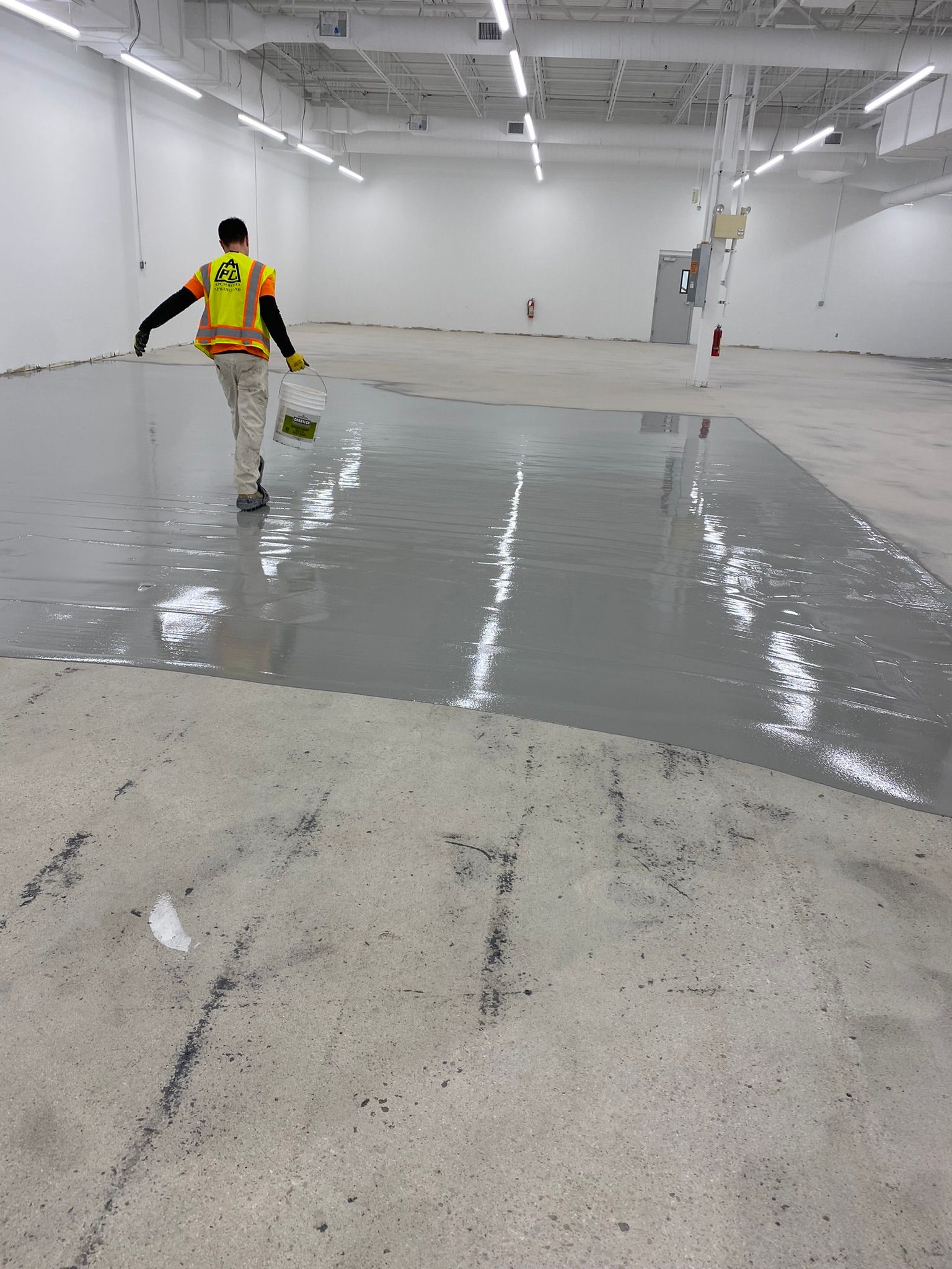 Worker pouring or smoothing gray floor coating in a large empty warehouse interior