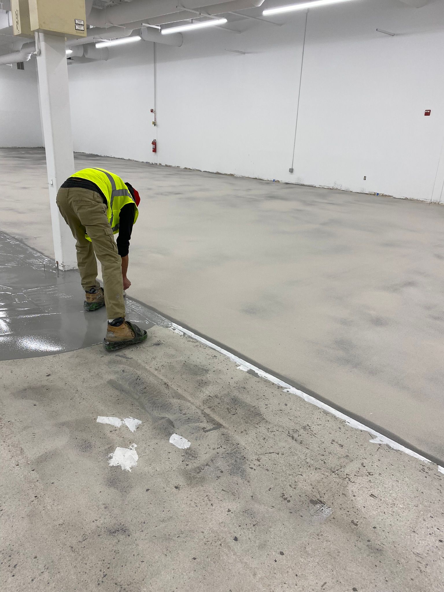 Worker in a yellow safety vest bending over a concrete floor in a large empty industrial space.
