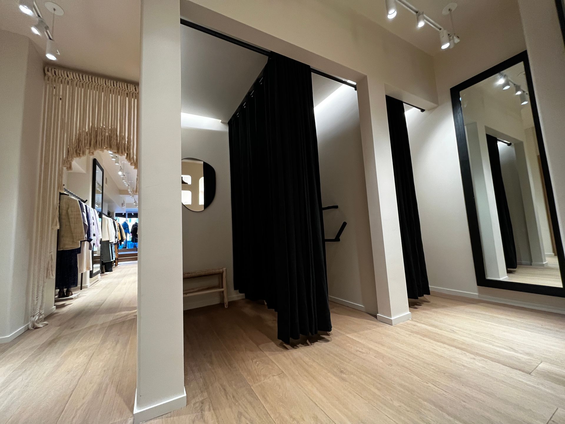 Boutique fitting room with white walls, wood floors, black curtains, and a large mirror in a hallway-like space