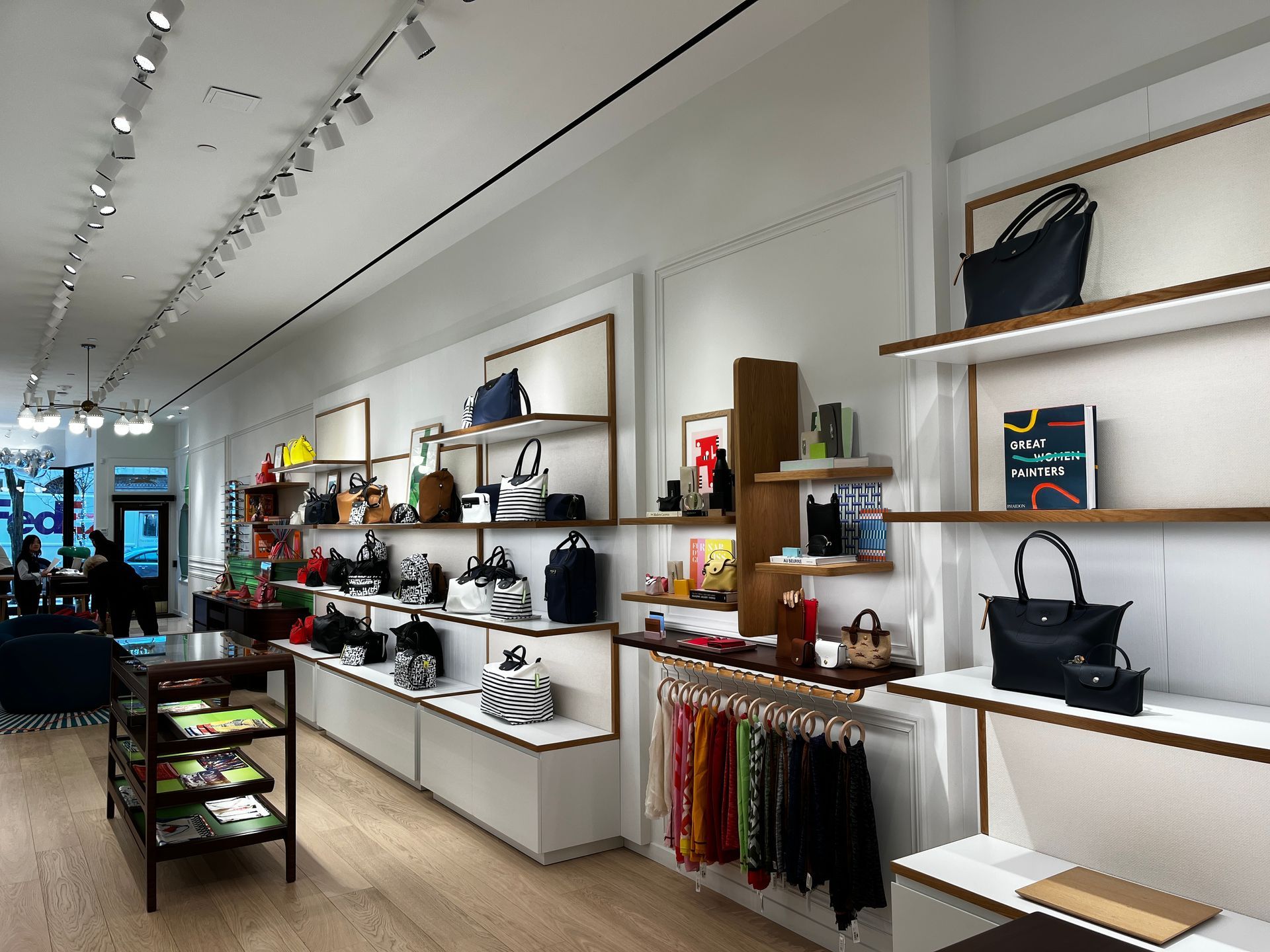 Bright boutique interior with white shelves, handbags, accessories, and clothing displays along a long aisle.