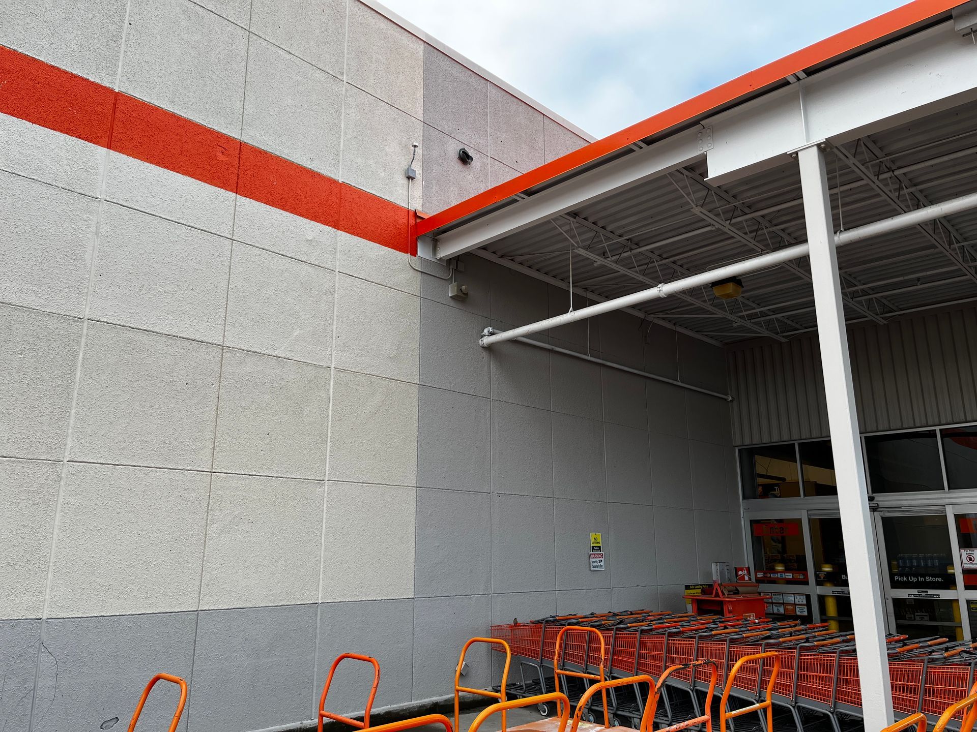 Orange shopping carts outside a store entrance under a white overhang with a gray wall and orange stripe