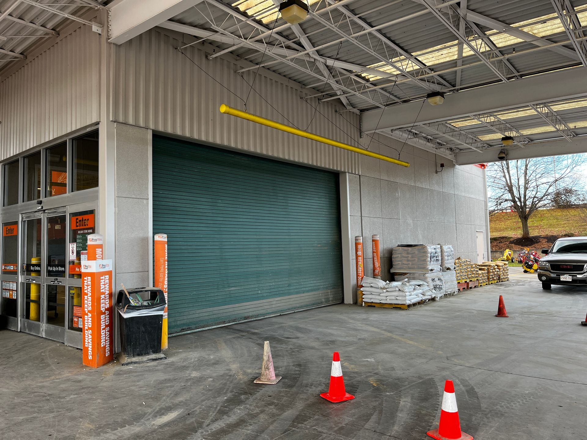 Warehouse loading dock with closed green roll-up door, orange cones, and stacked pallets under an overhang.