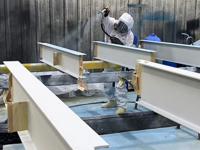 Worker spray-painting white steel beams in an industrial workshop