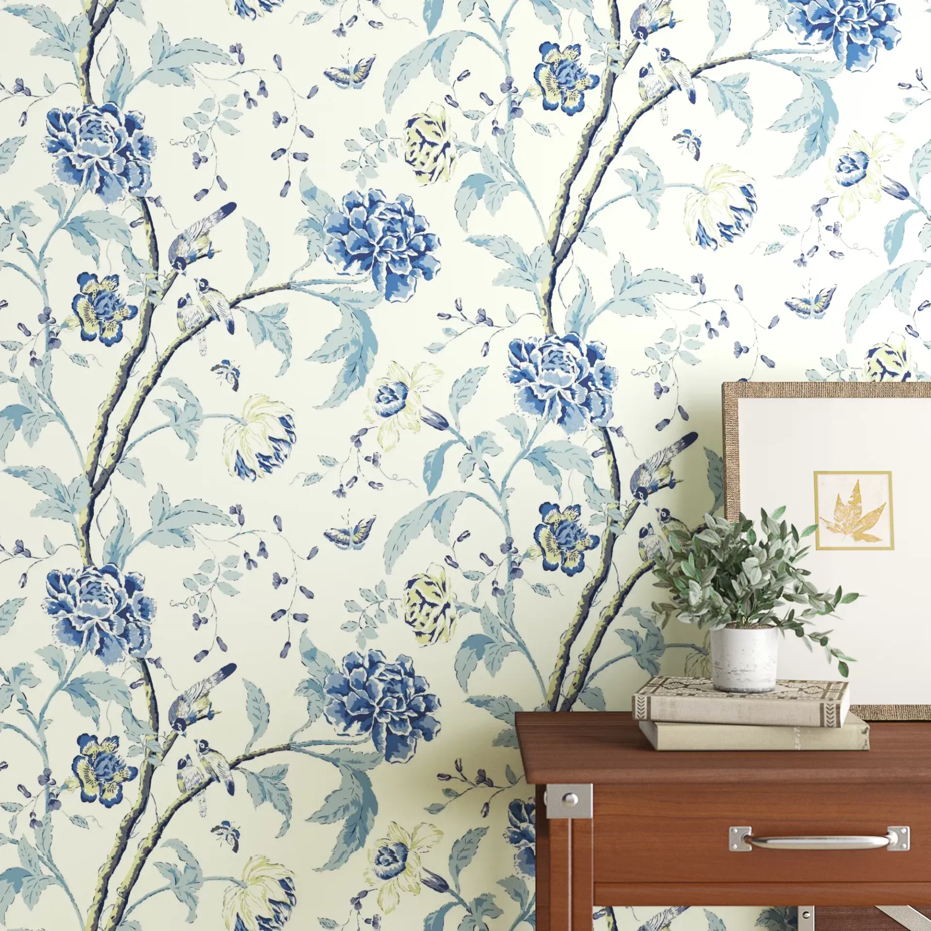 Blue floral wallpaper above a wooden dresser with a small plant and framed art.