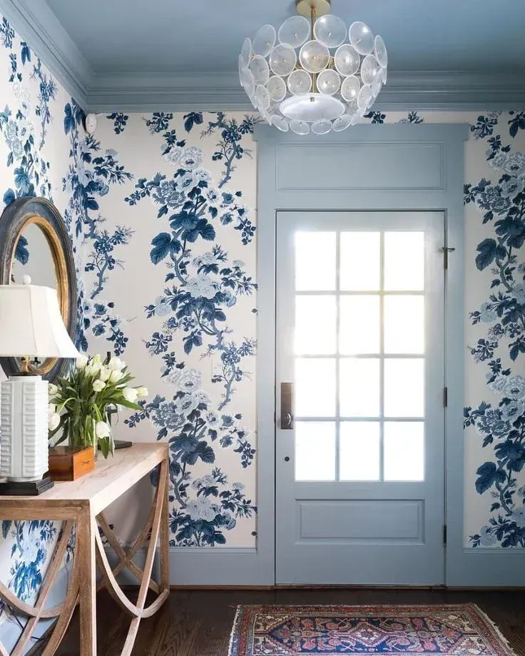 Blue floral wallpapered entryway with a white door, chandelier, and console table with mirror and lamp