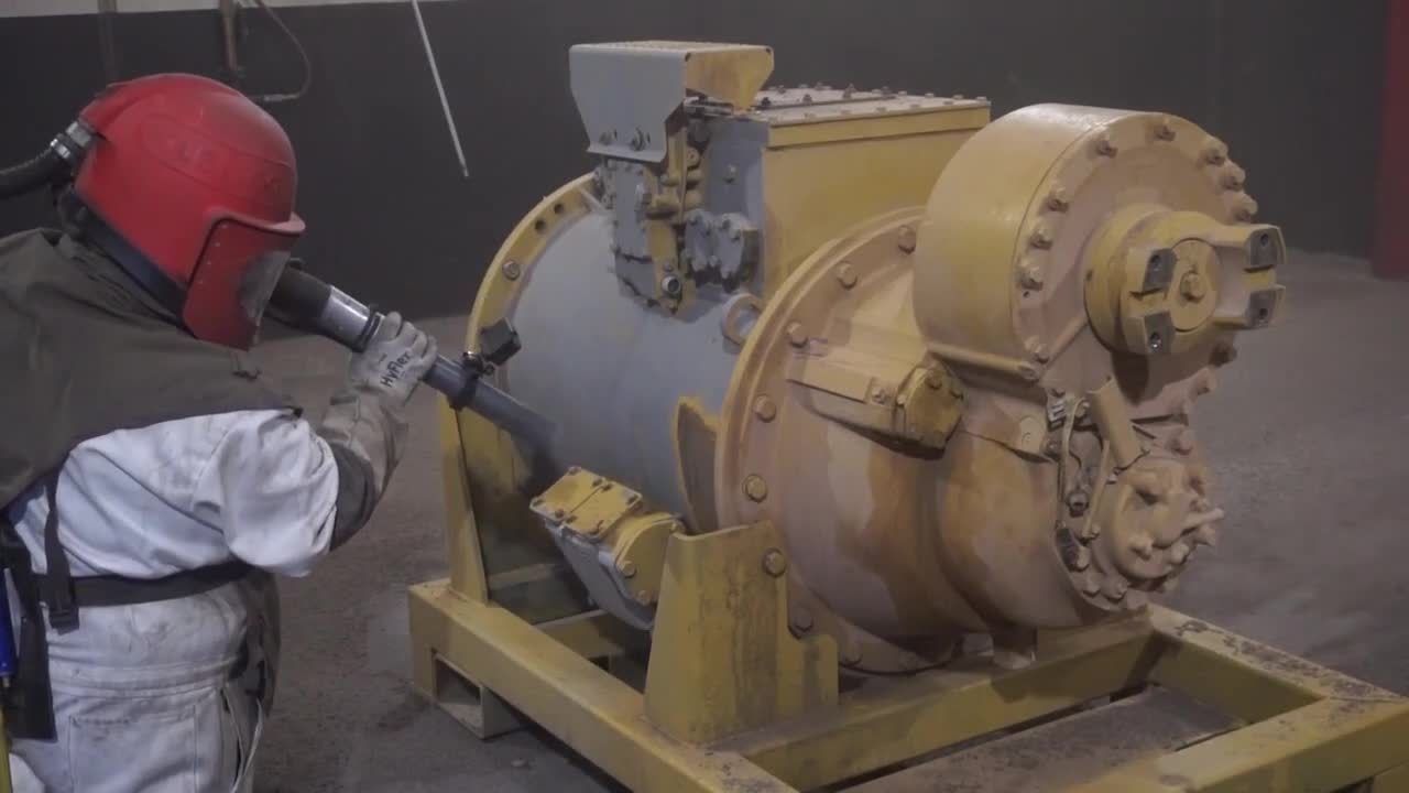 Worker in red helmet spraying a large industrial machine with paint or coating.
