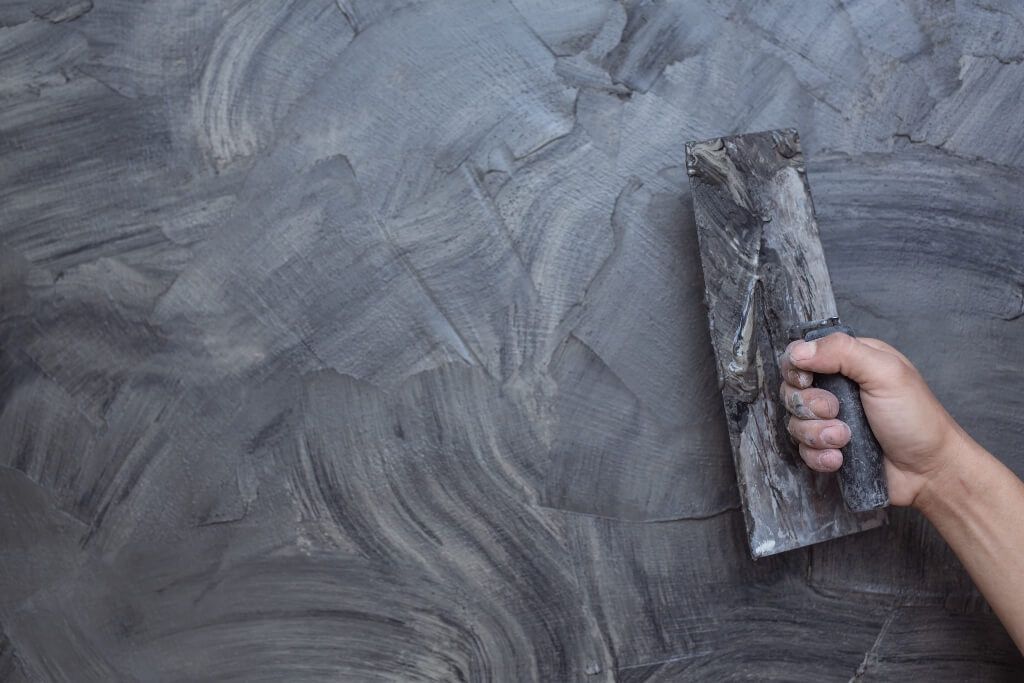 Hand holding a putty knife smoothing gray plaster on a wall