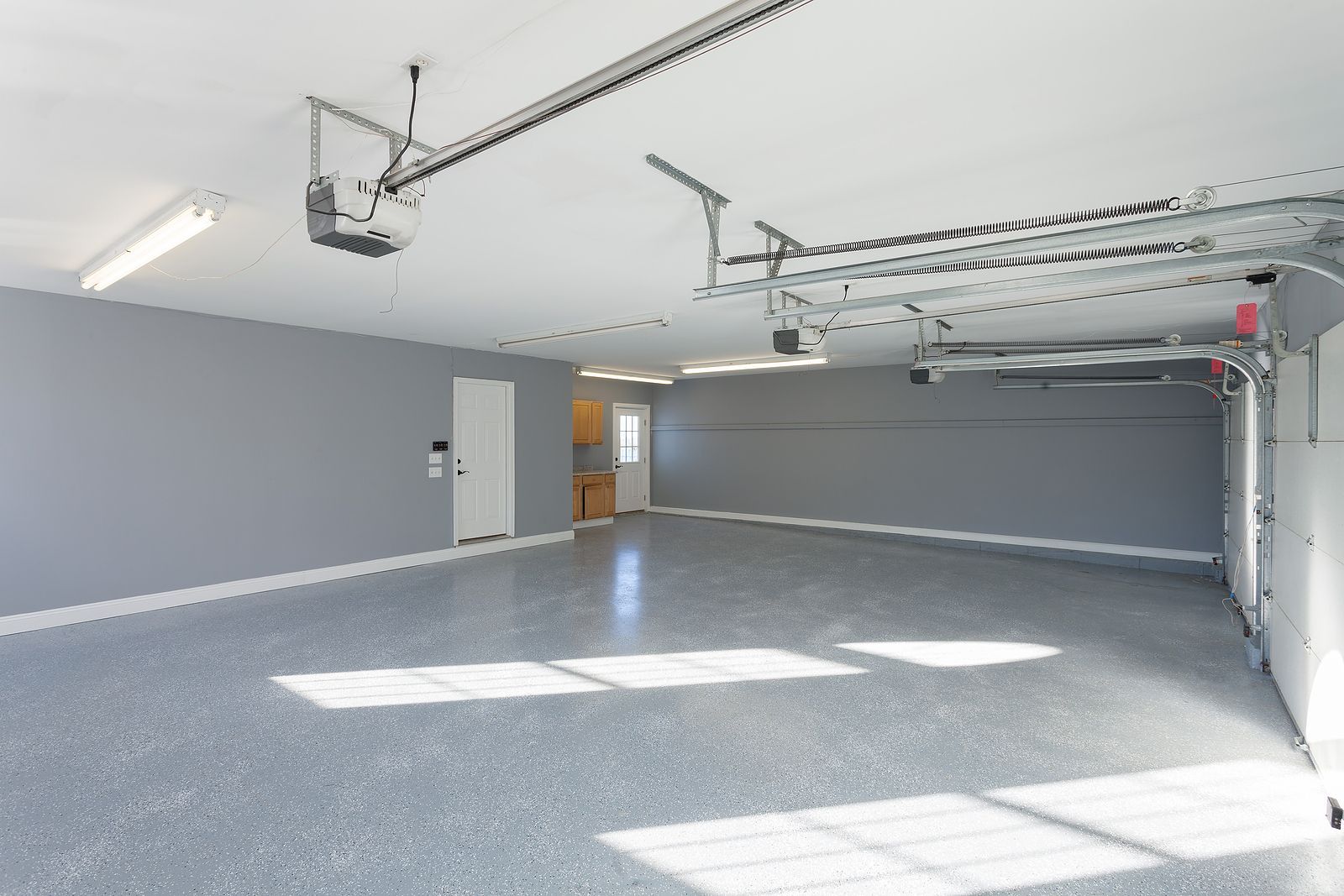 Empty gray garage with open door, concrete floor, ceiling-mounted opener, and bright sunlight on the floor