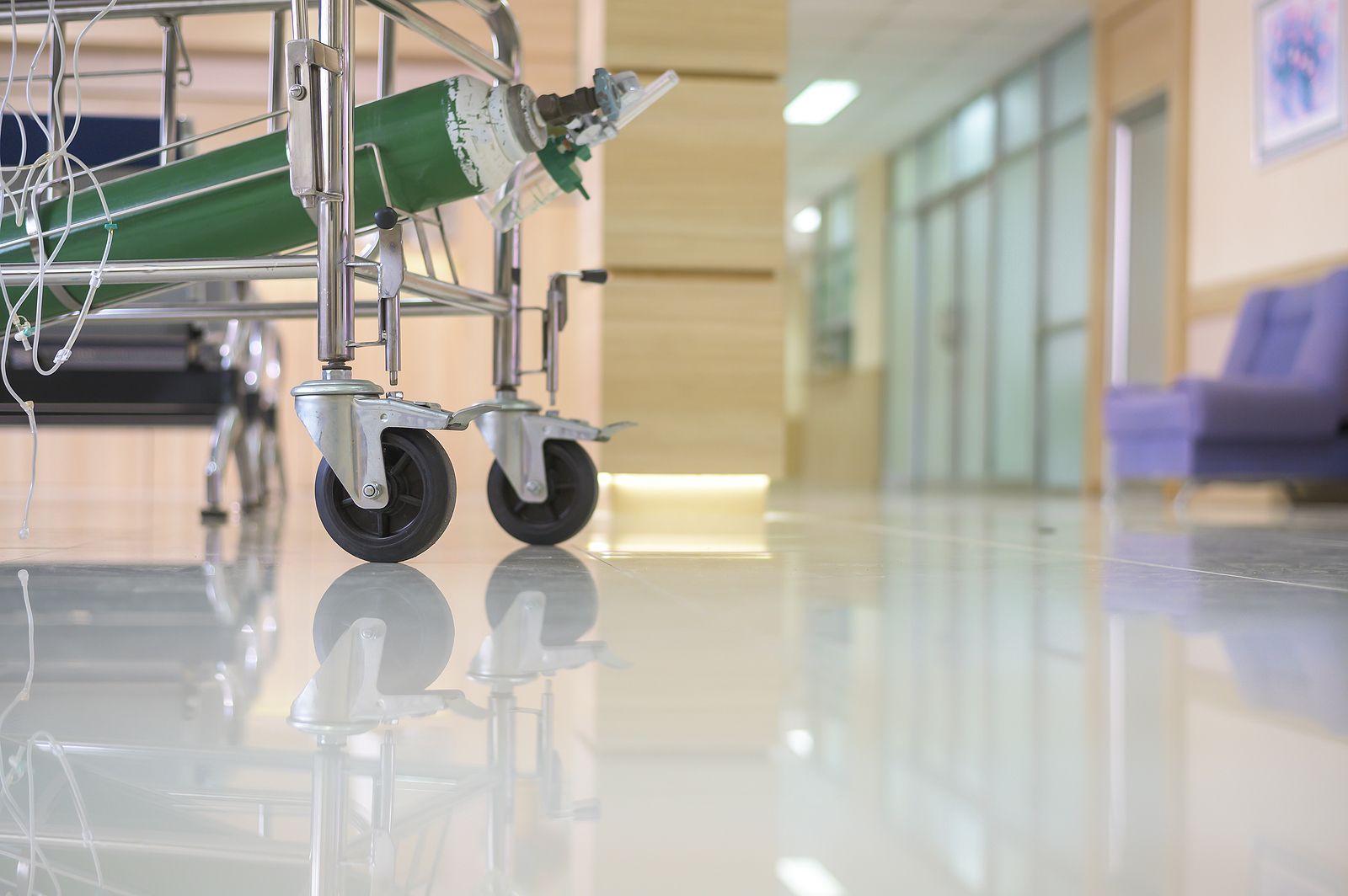 Hospital gurney with green side rail in a bright hallway, reflected on a polished floor.