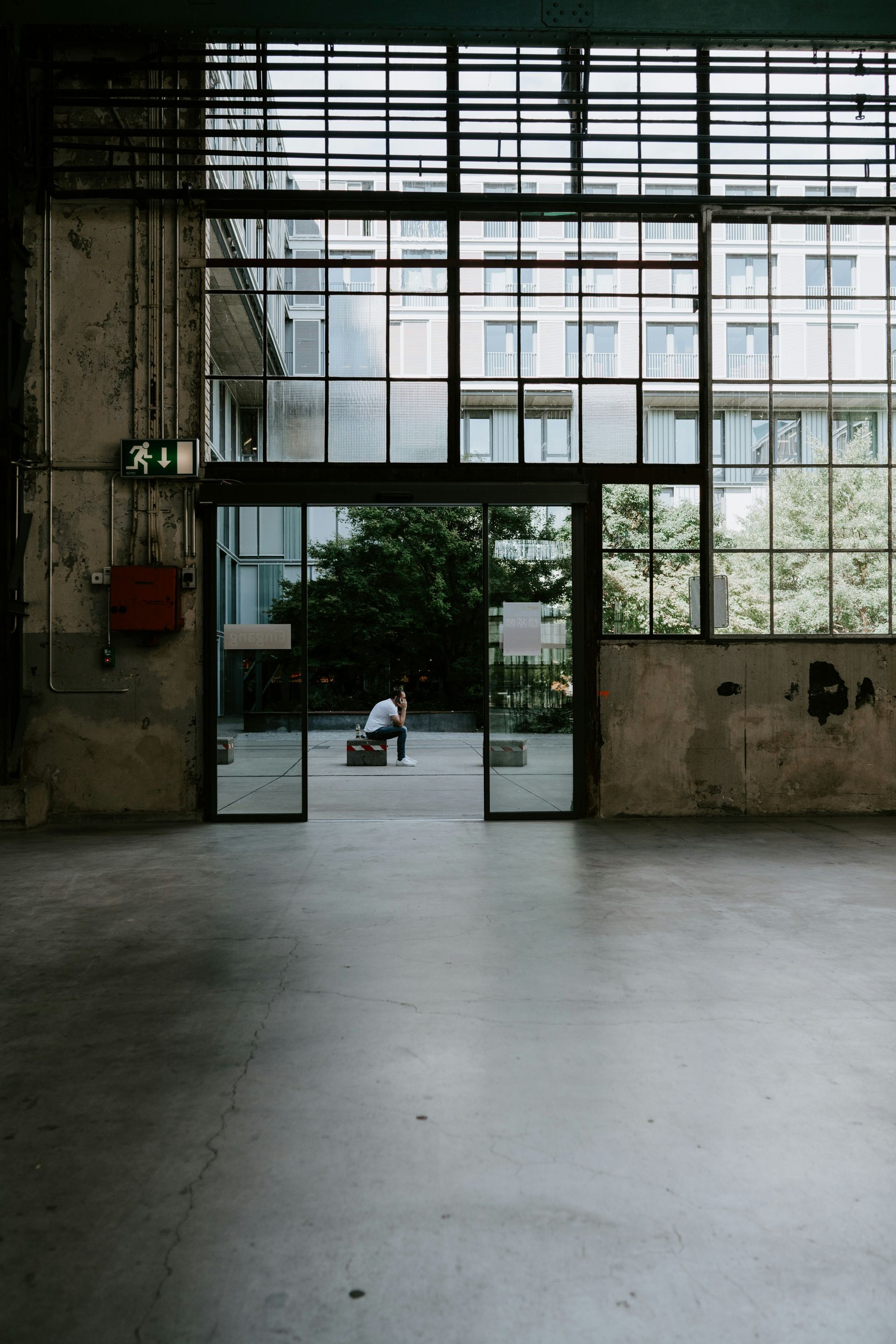 Empty industrial hallway with a glass doorway opening to a courtyard and a person in the distance.