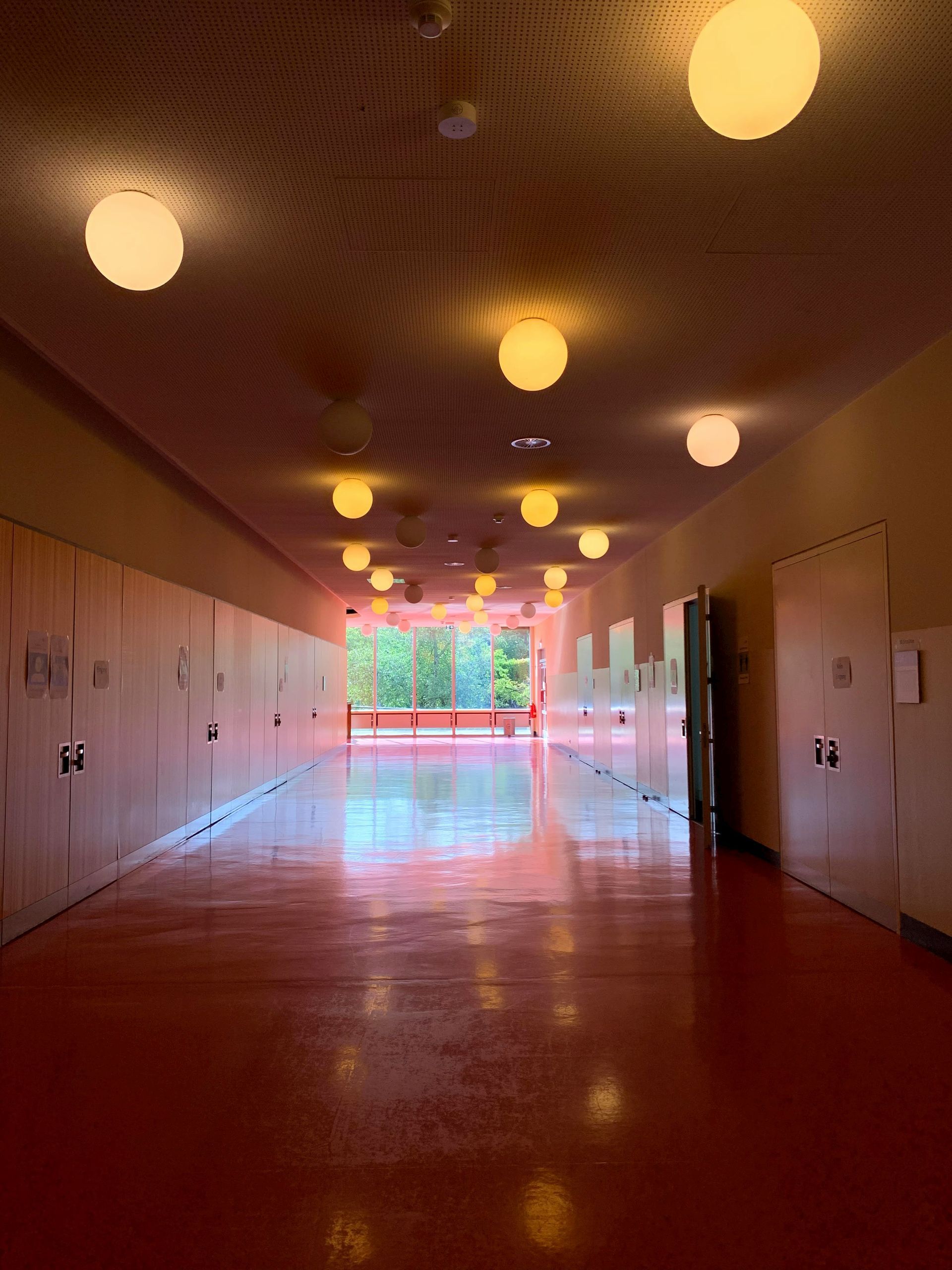 Empty hallway with pink walls, hanging round lights, and a glossy floor leading to bright windows.