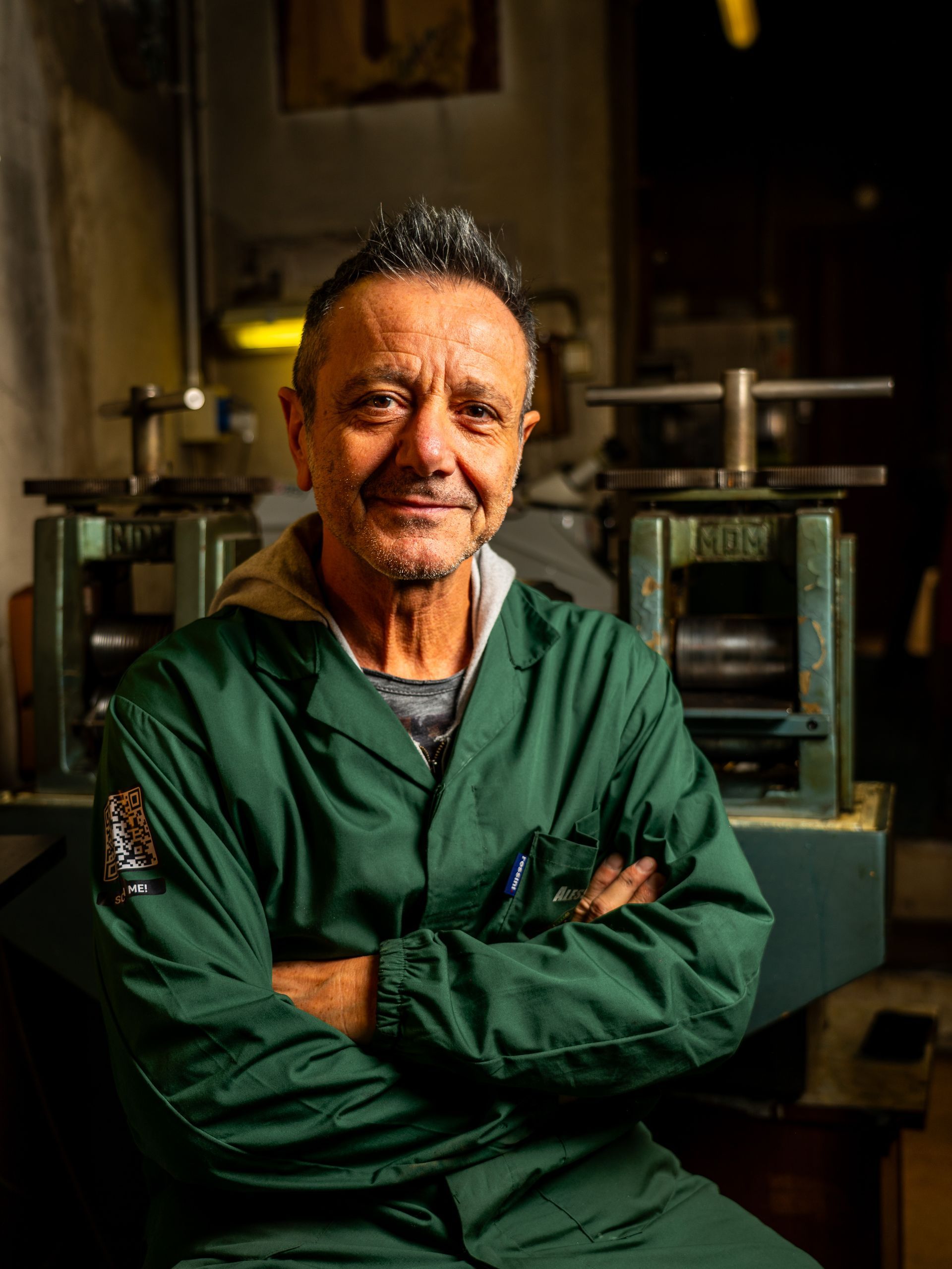 Una persona in uniforme da lavoro verde, con le braccia incrociate, in piedi in un'officina con attrezzature industriali sullo sfondo.