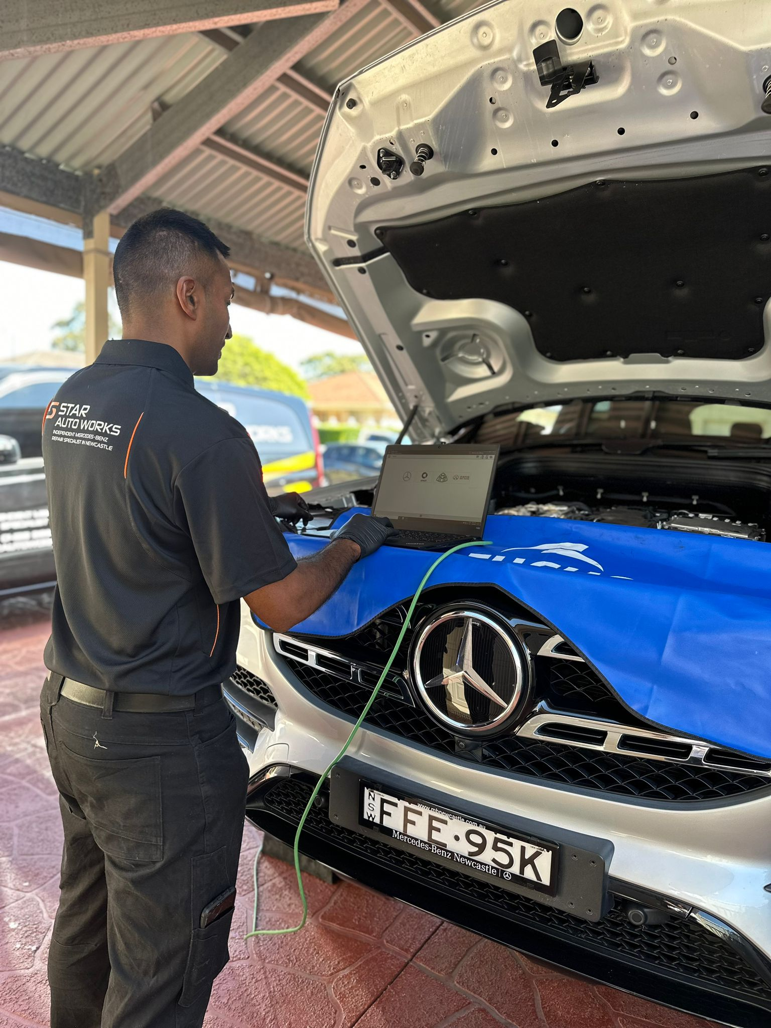 A technician in a black uniform uses a diagnostic laptop on a silver Mercedes-Benz with the hood open under a carport.