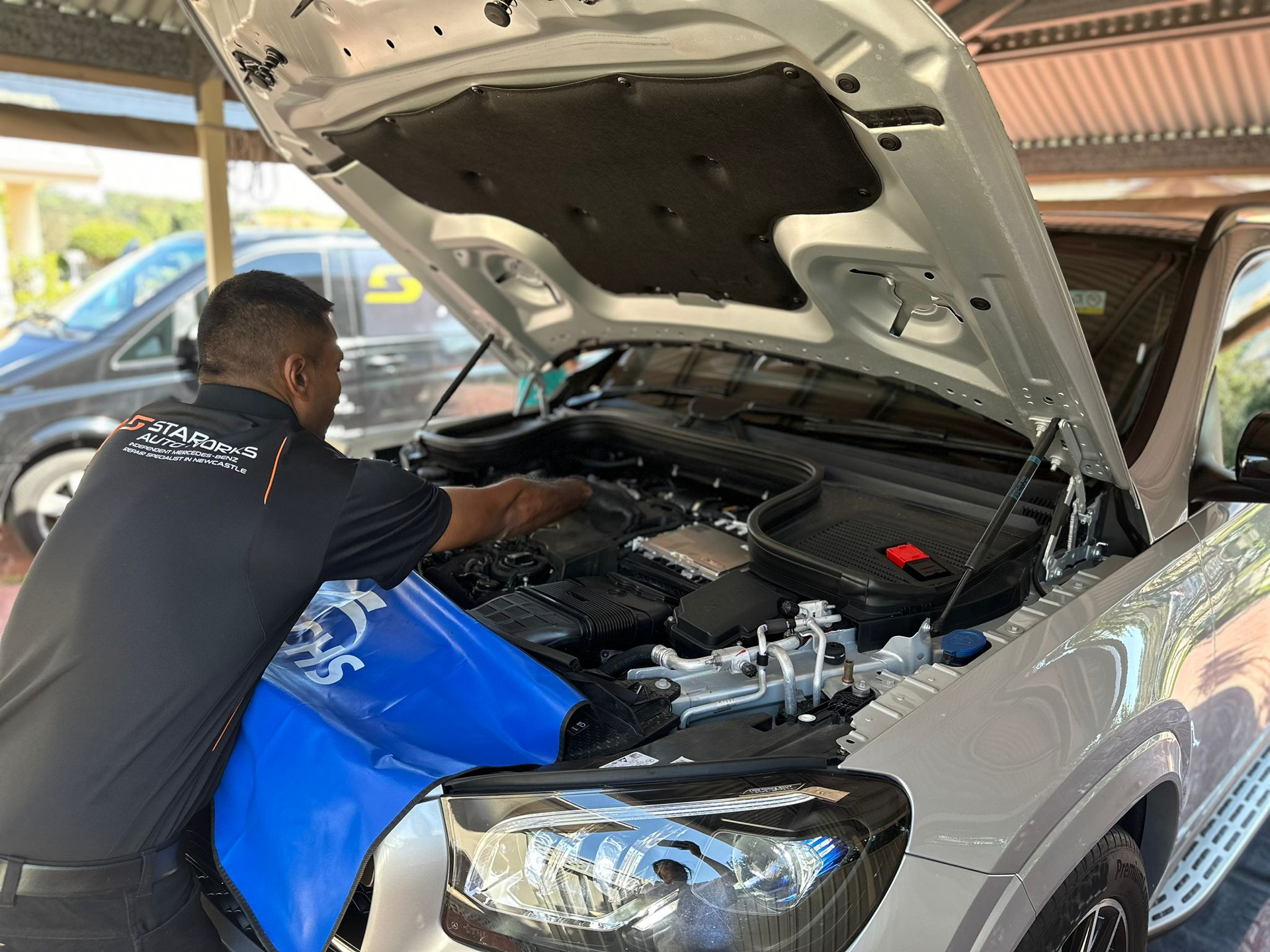 A technician in a black uniform works on the open engine bay of a silver SUV under a covered structure.