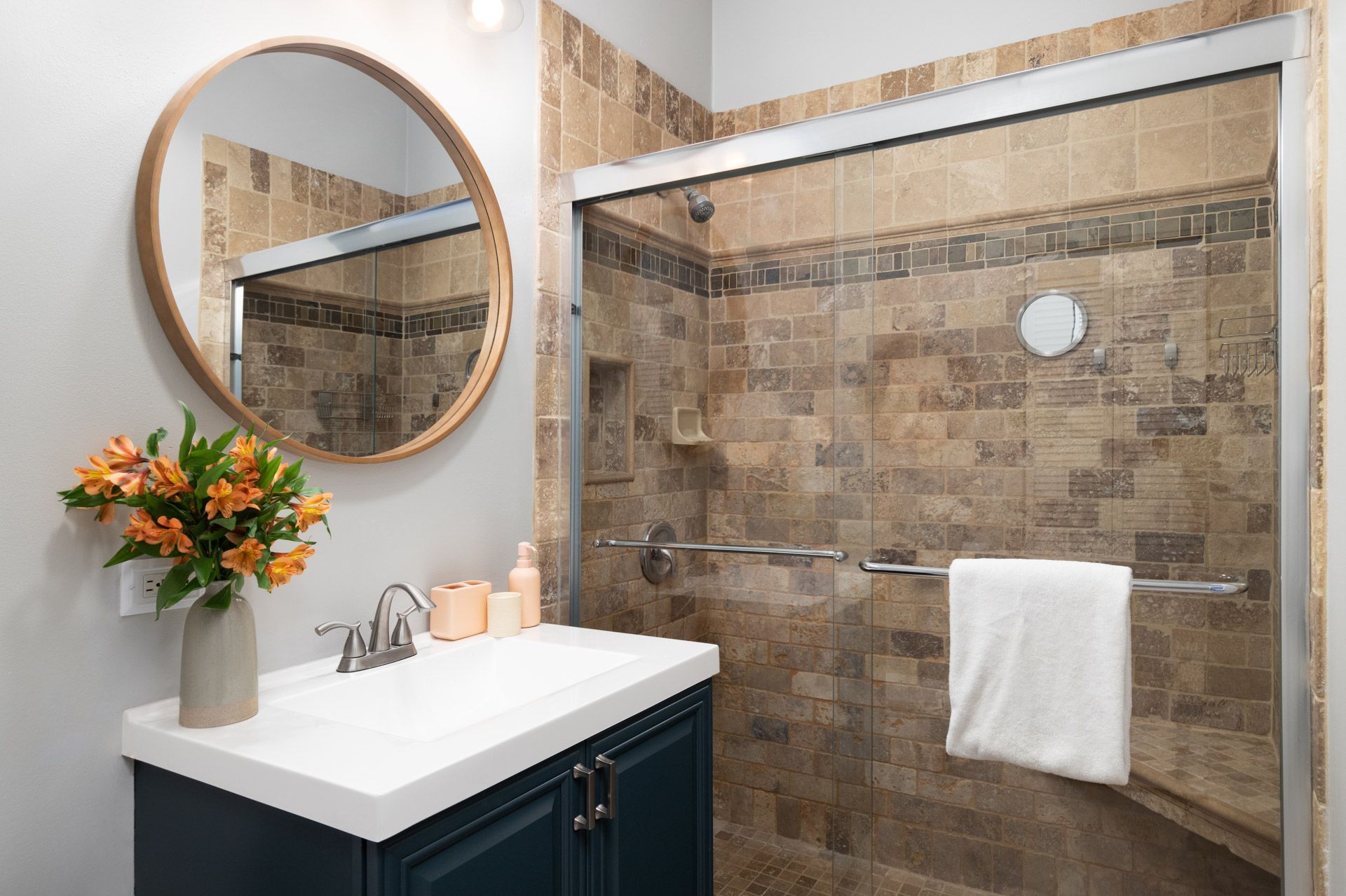 Modern bathroom with round mirror, white vanity, and glass shower with beige tile