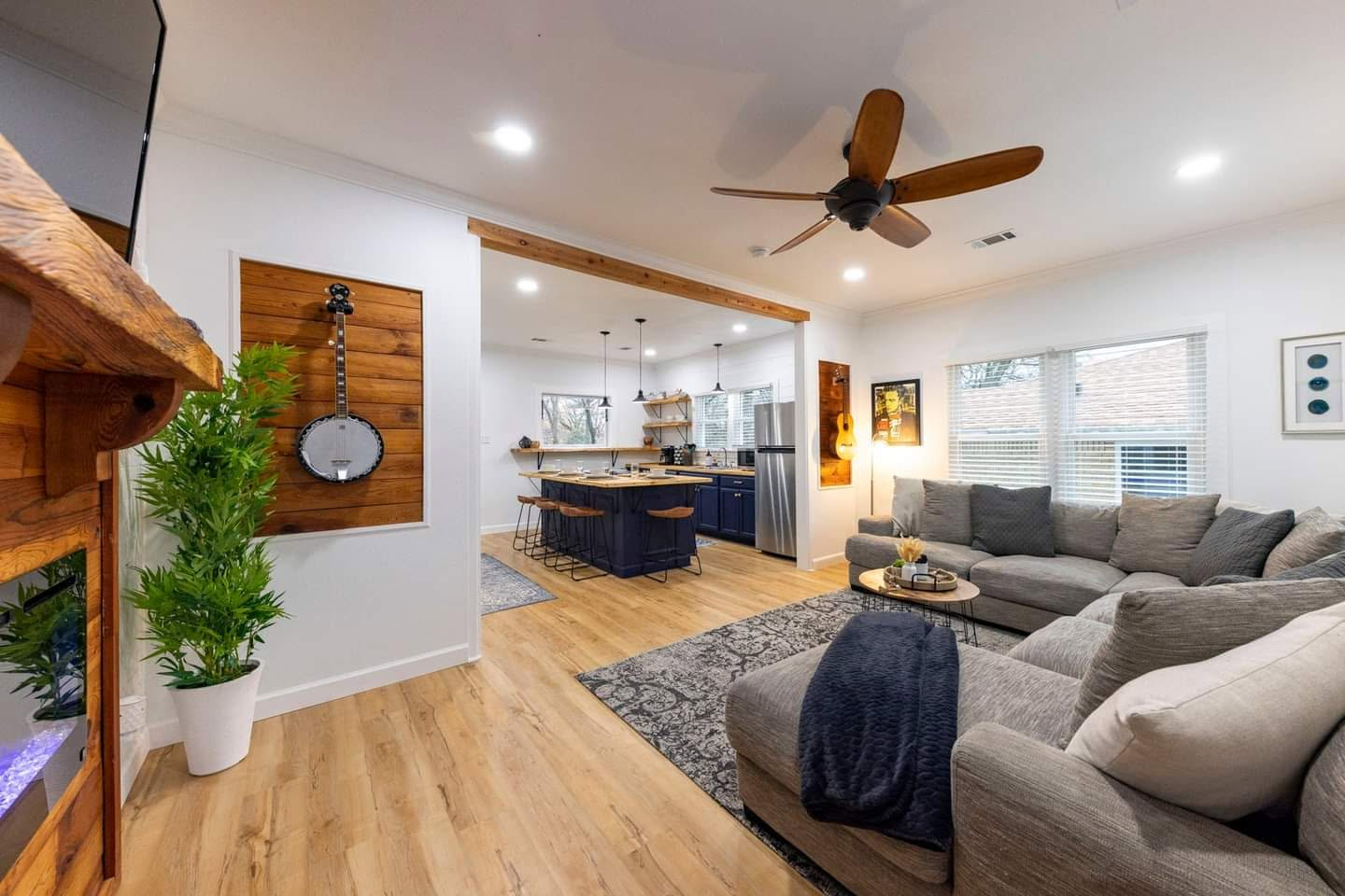 Bright open living room with gray sectional, wood floors, ceiling fan, and kitchen in the background