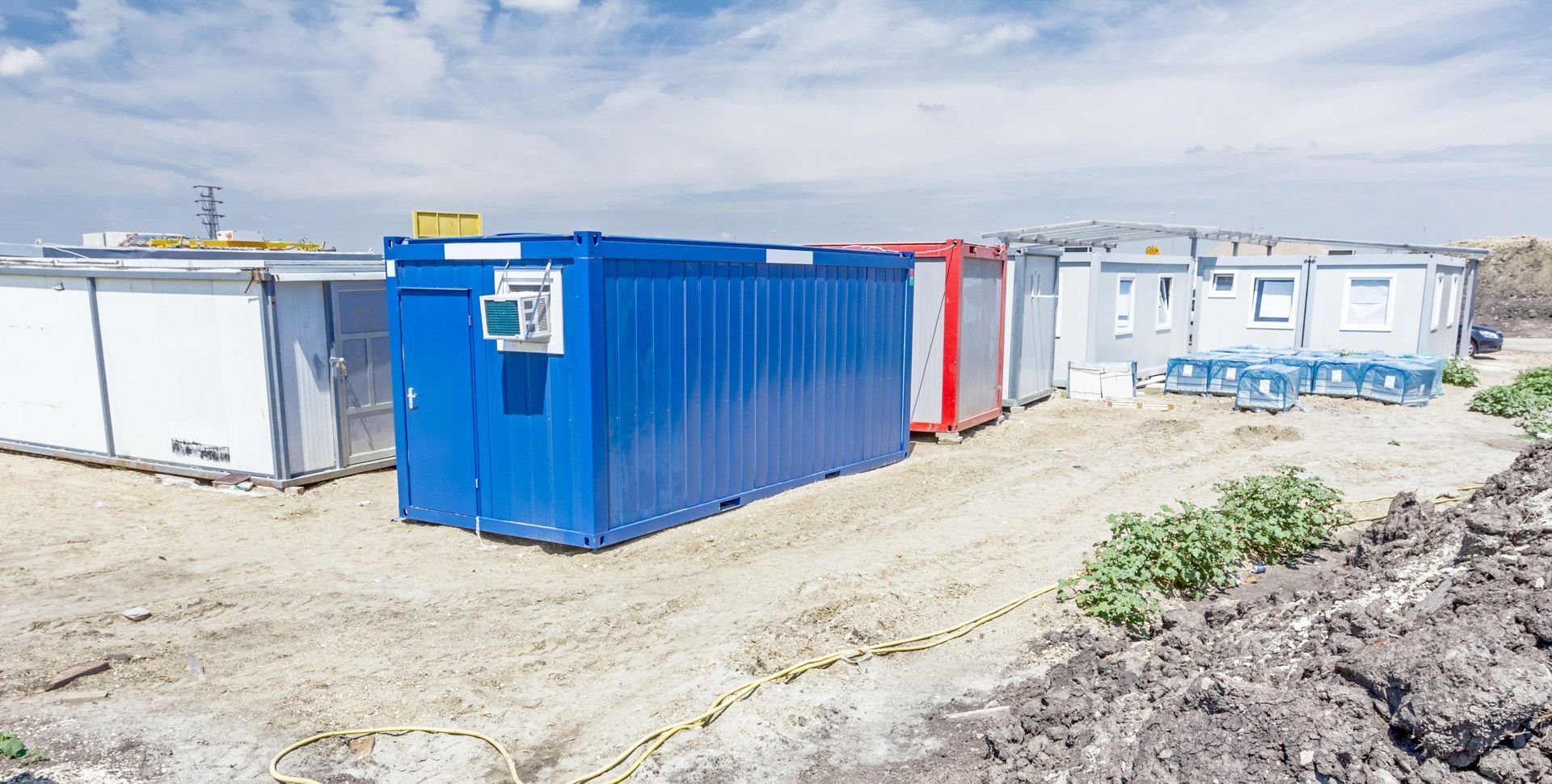 Blue and white shipping containers on a sandy construction site under a bright sky