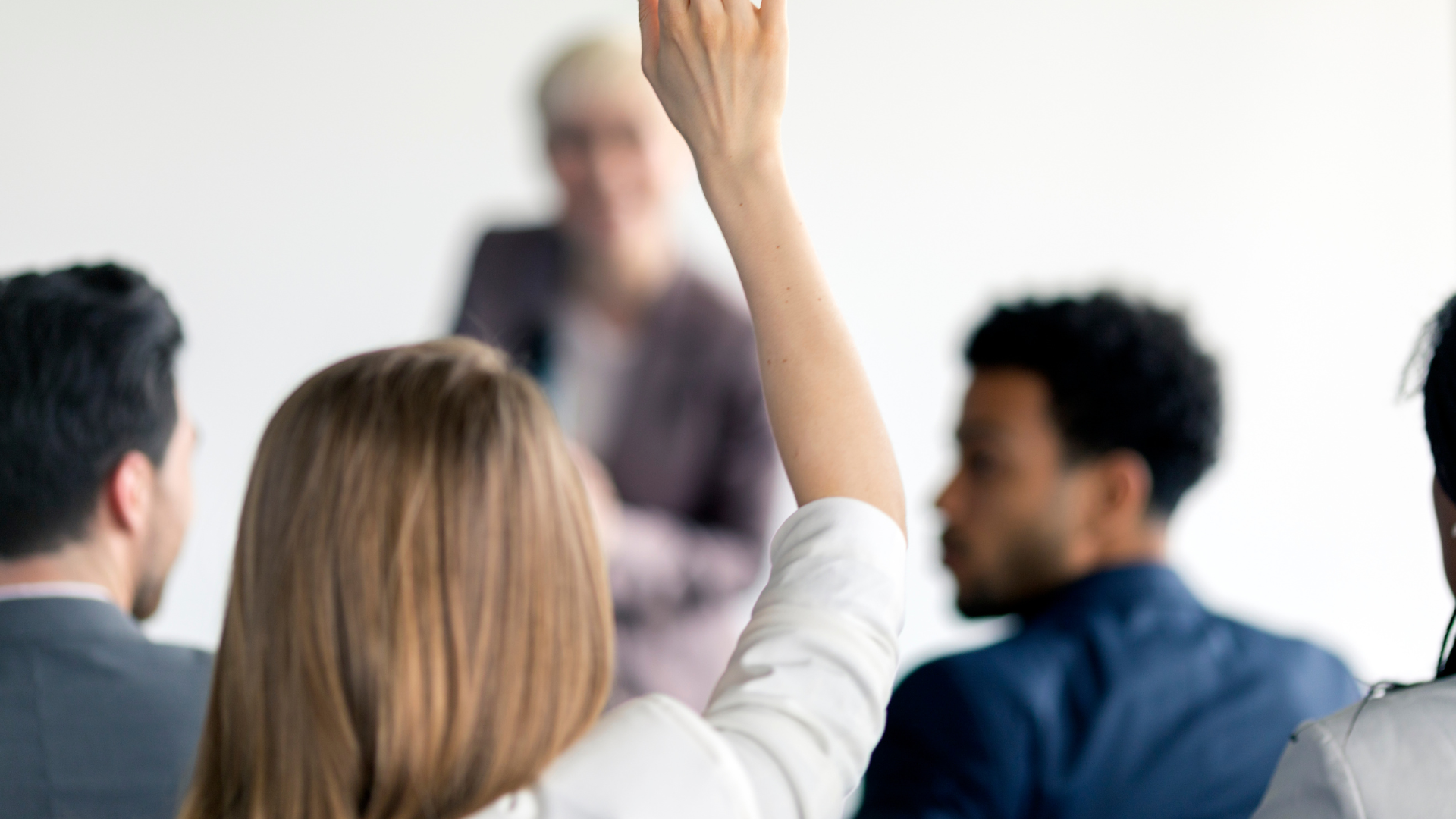 Audience member raises hand in a blurred meeting room while others listen to a speaker at the front