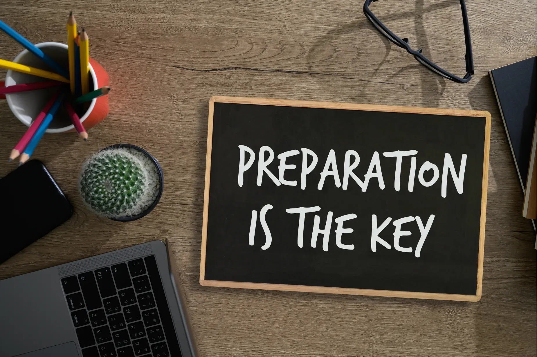 Desk with chalkboard reading “Preparation is the key,” surrounded by office supplies and a laptop.