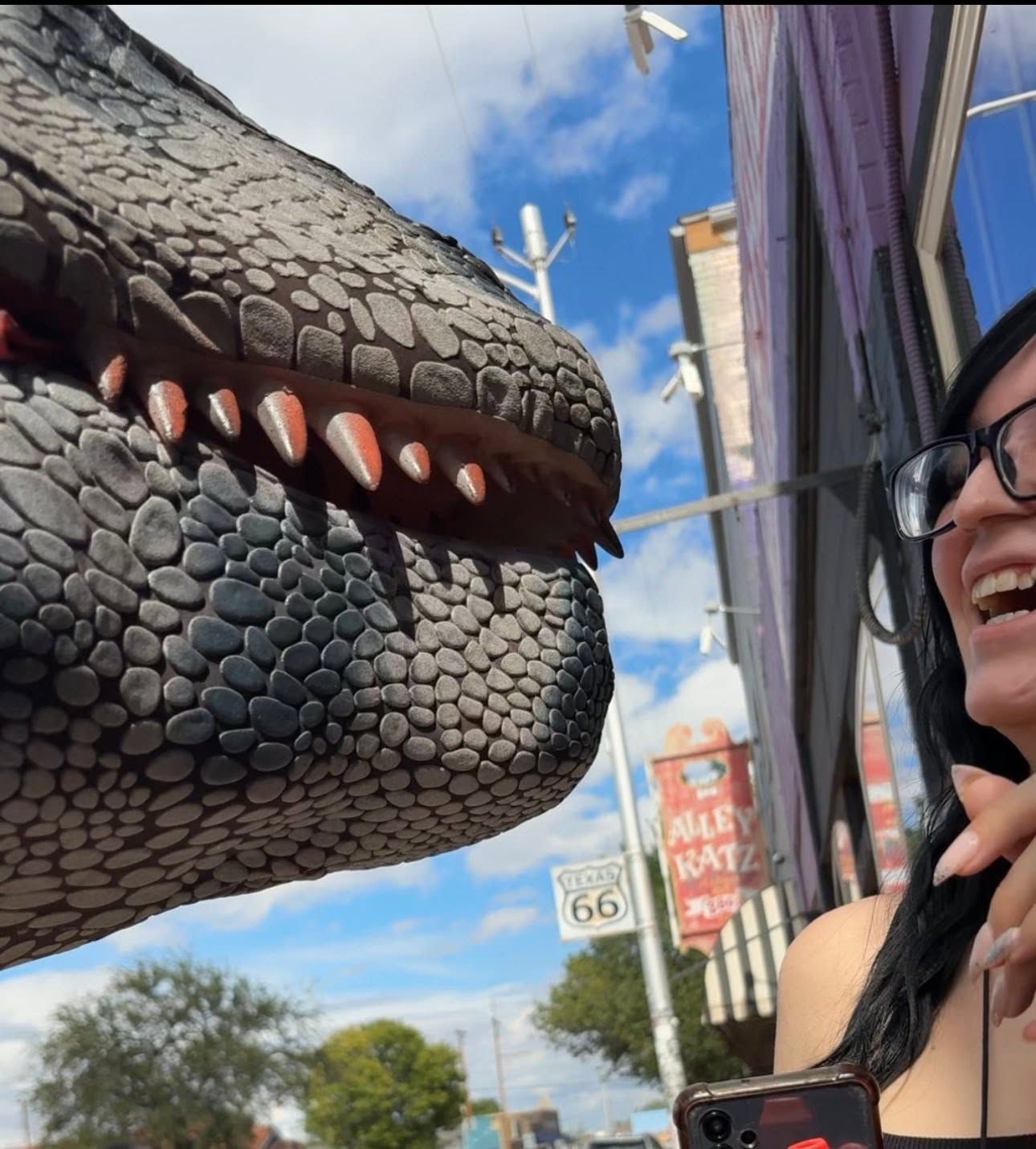 A large dragon head sculpture faces a smiling person wearing glasses on a sunny street.