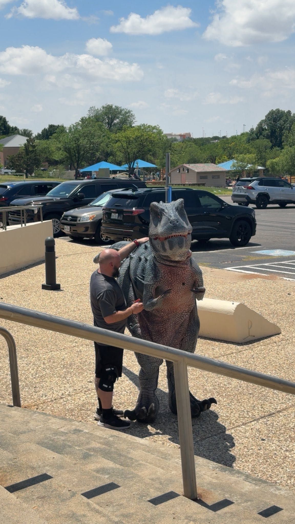 Person in a camouflage costume stands outdoors near a railing, with a child holding onto them in a parking lot.
