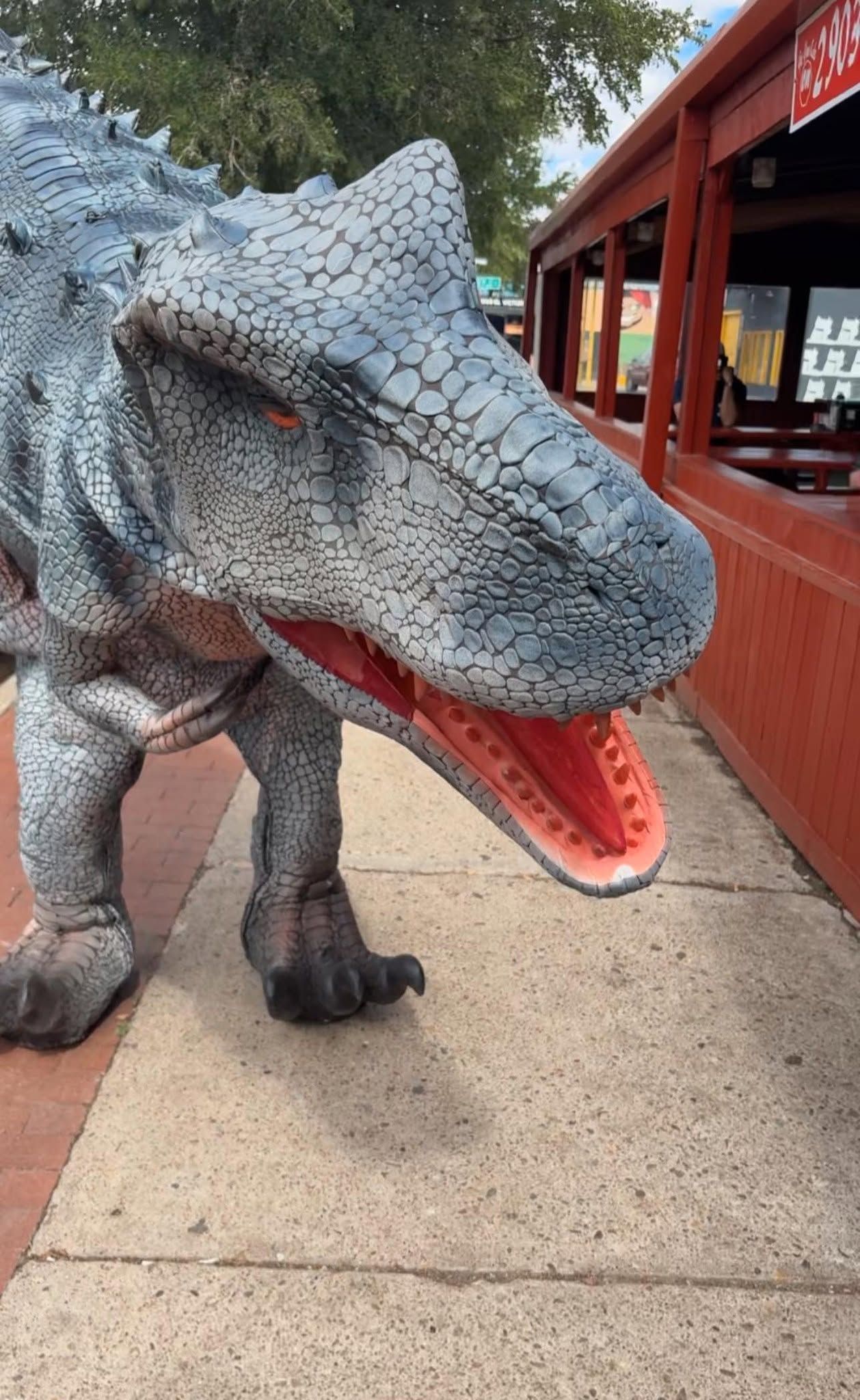 Large gray dinosaur statue with open red mouth beside a wooden walkway outdoors