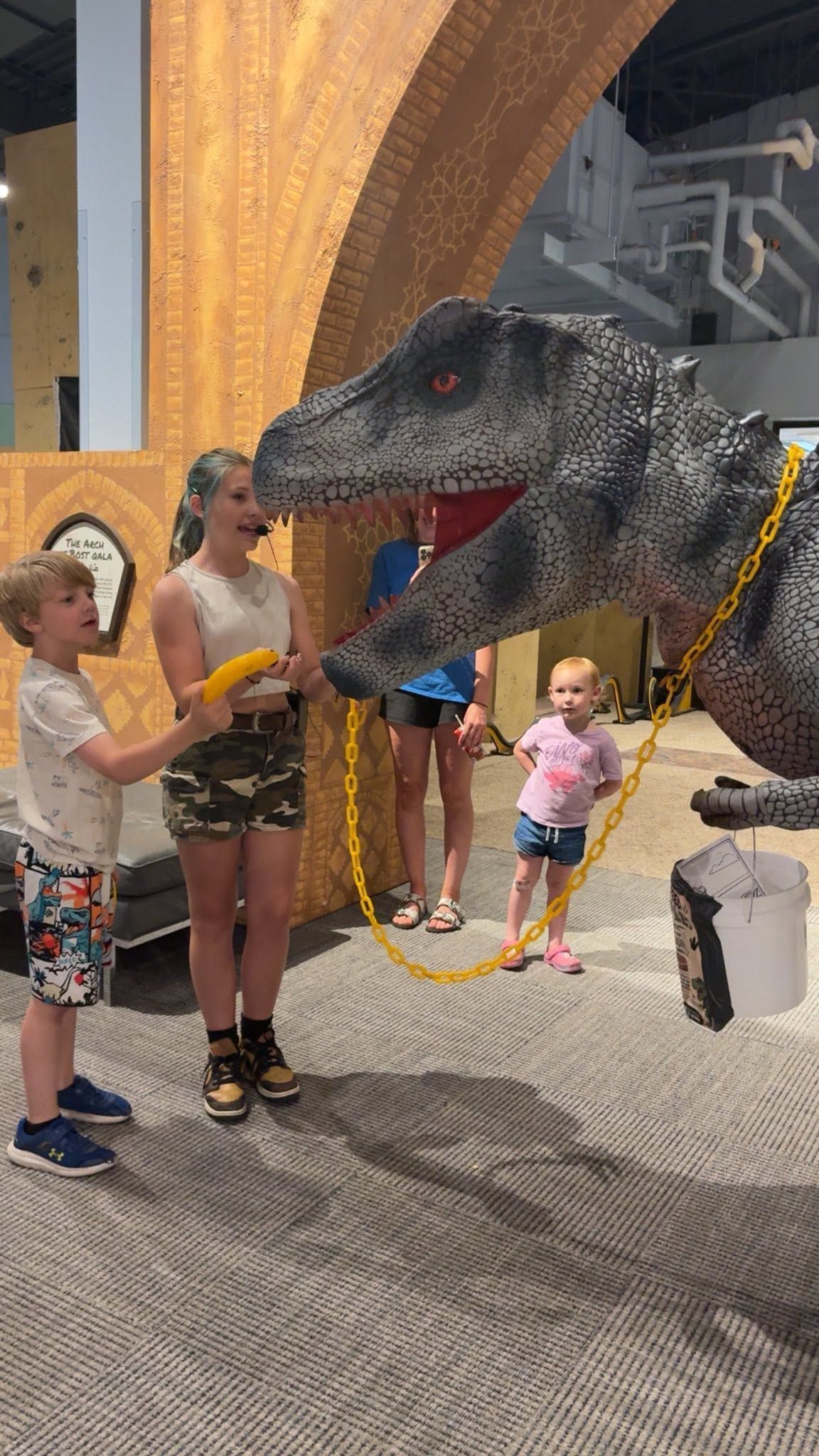 Child posing beside a large dinosaur sculpture in a museum or exhibit hall, with rope barrier and visitors nearby