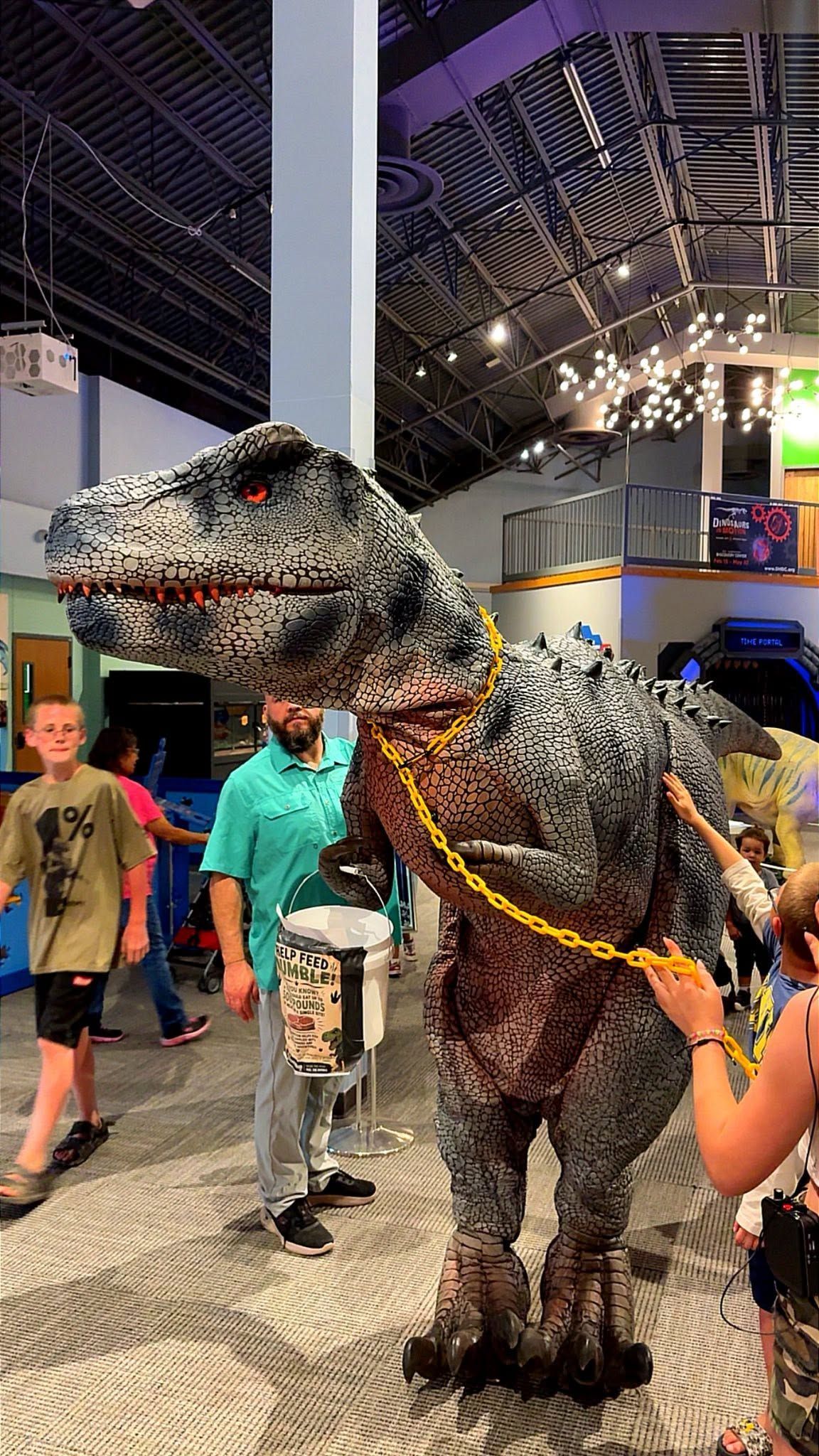 Person in dinosaur costume at a busy indoor event hall, with children nearby and bright lights overhead