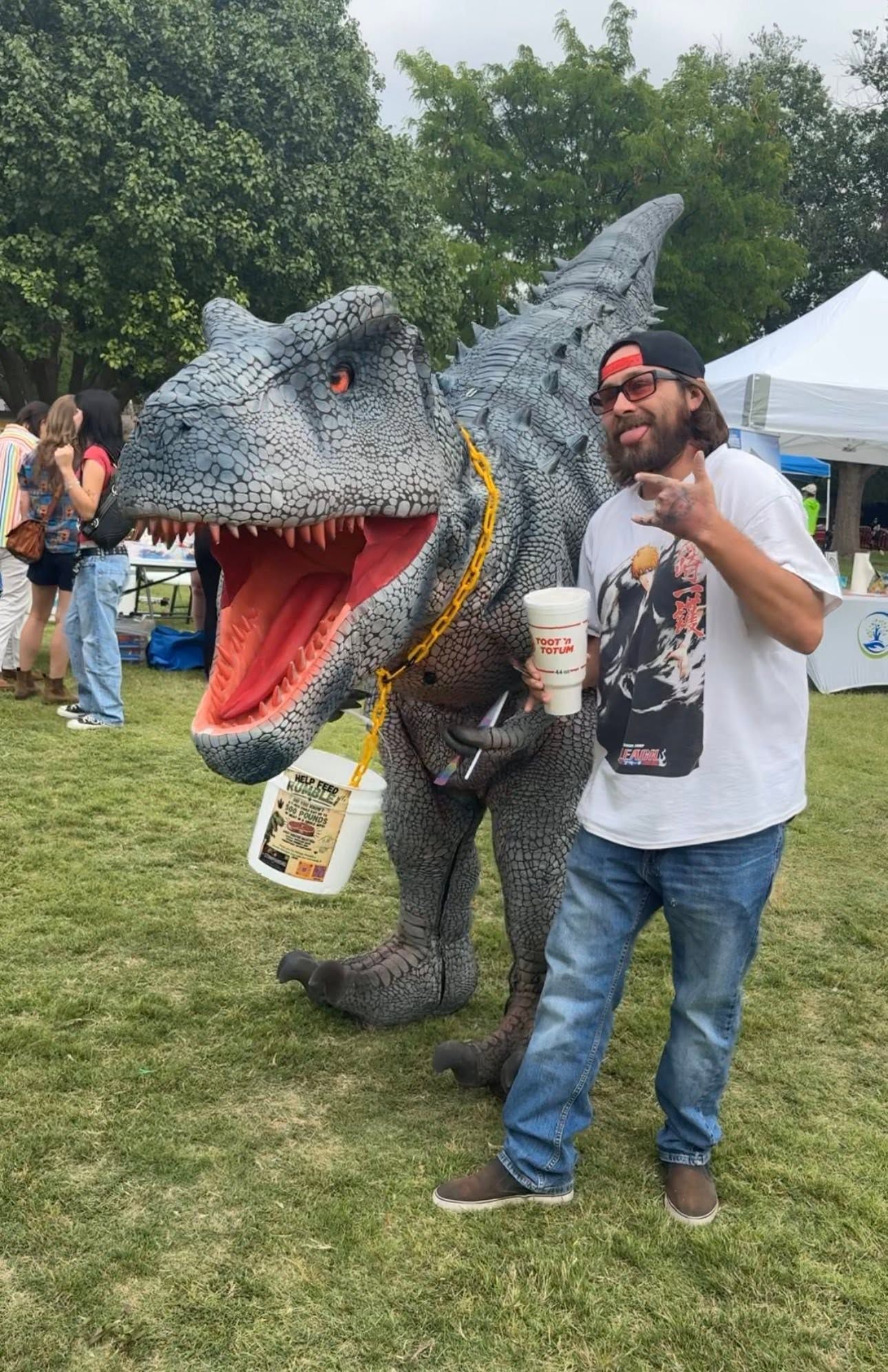 Man posing beside a life-size T. rex costume in a park, holding a drink and smiling.