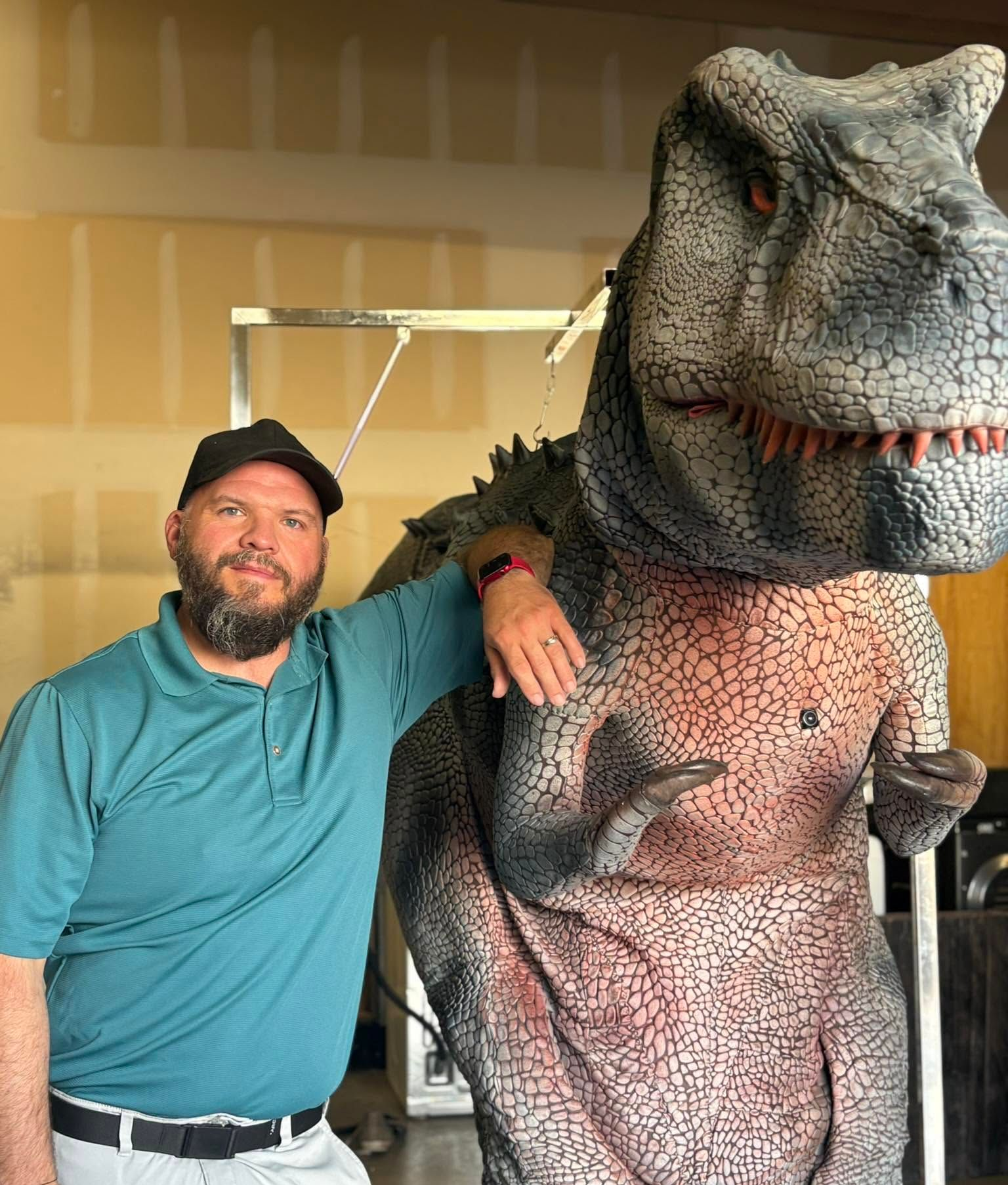 Man posing arm-in-arm with a life-size T. rex dinosaur statue indoors