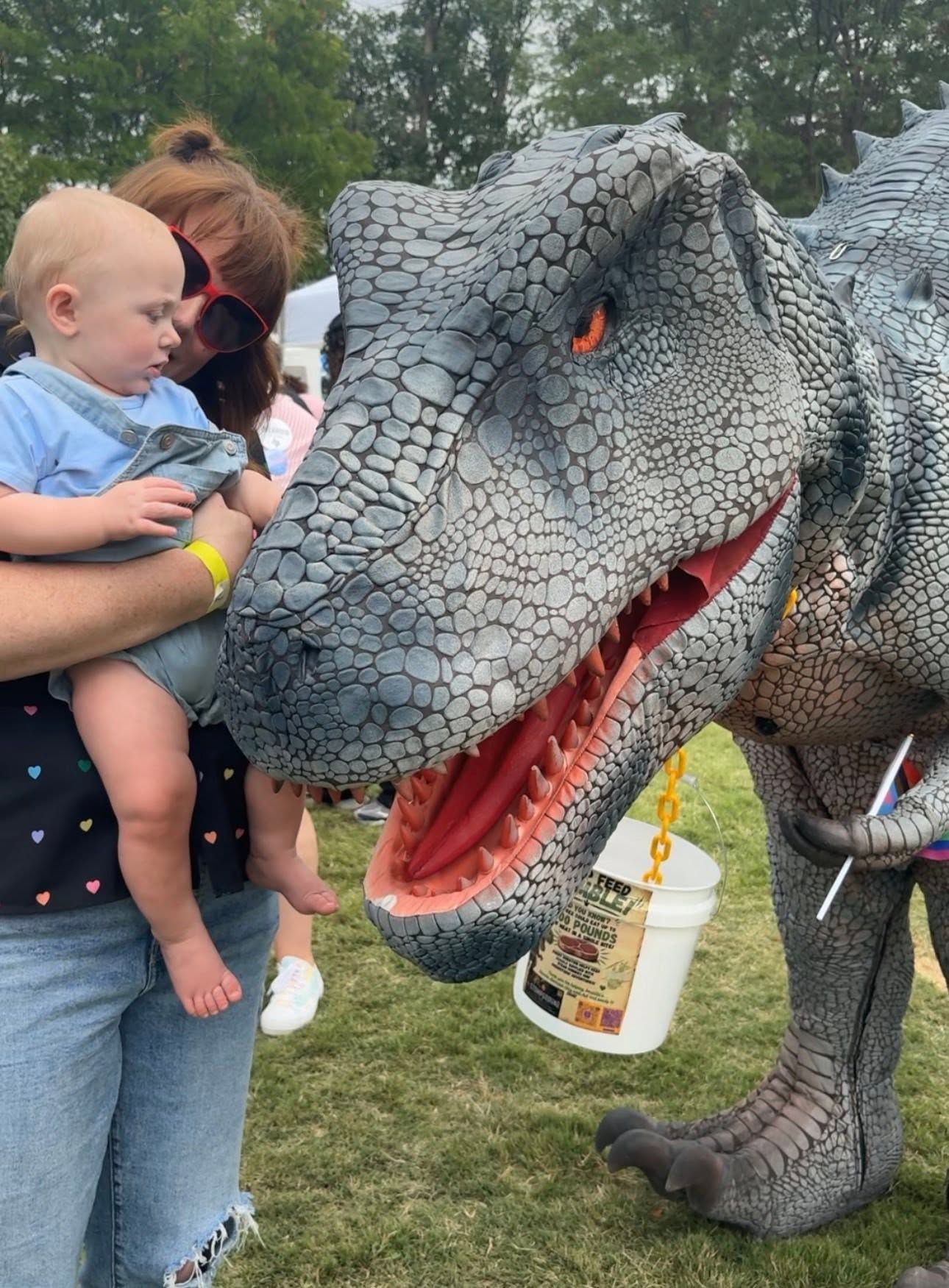 Woman holding baby near gray T. rex statue with open mouth at an outdoor event